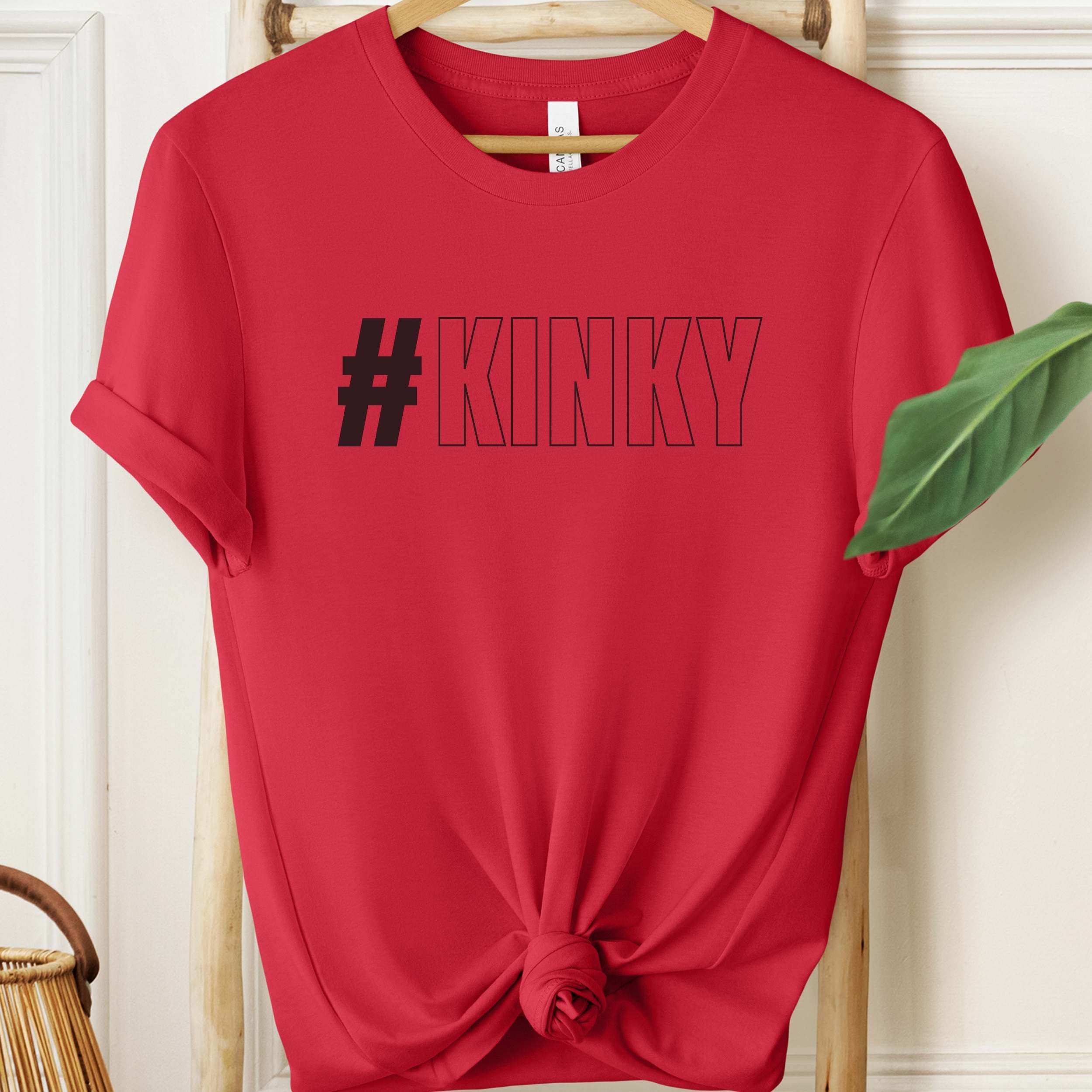 Red t-shirt with '#KINKY' text on a wooden hanger against a white wall.