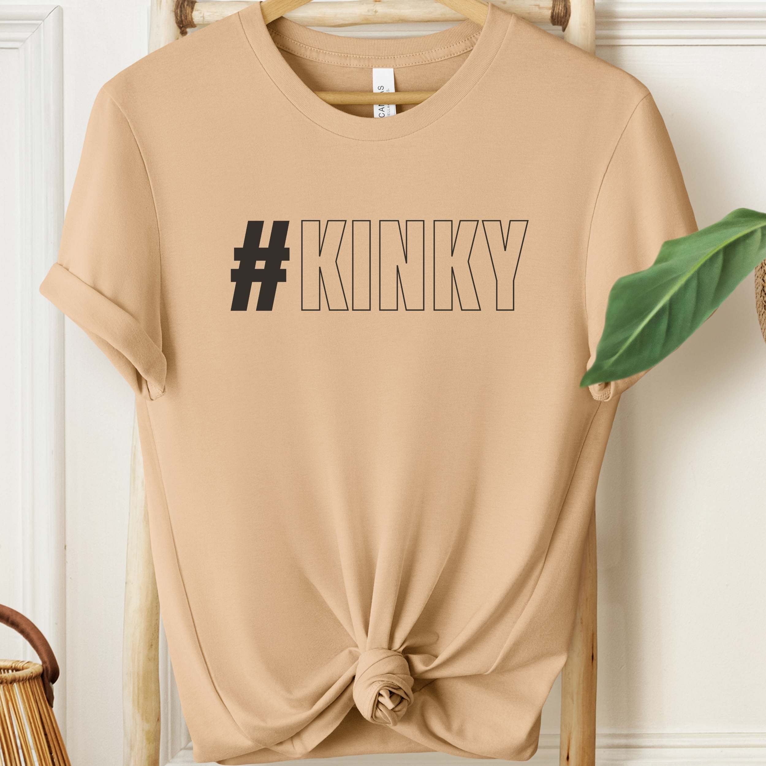 Beige t-shirt with '#KINKY' text on a wooden hanger against a white wall.