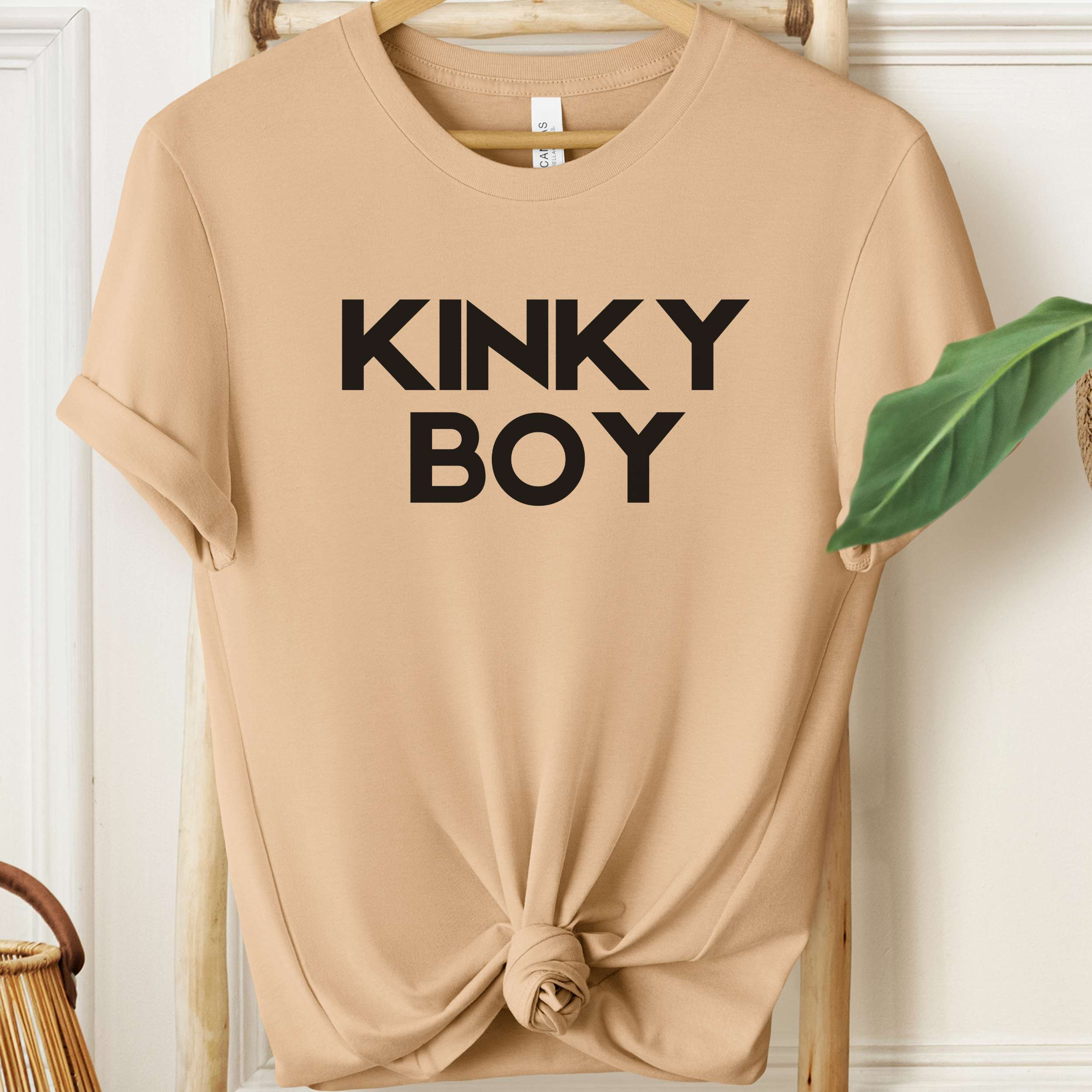 Beige t-shirt with 'KINKY BOY' text on a wooden hanger against a white wall.