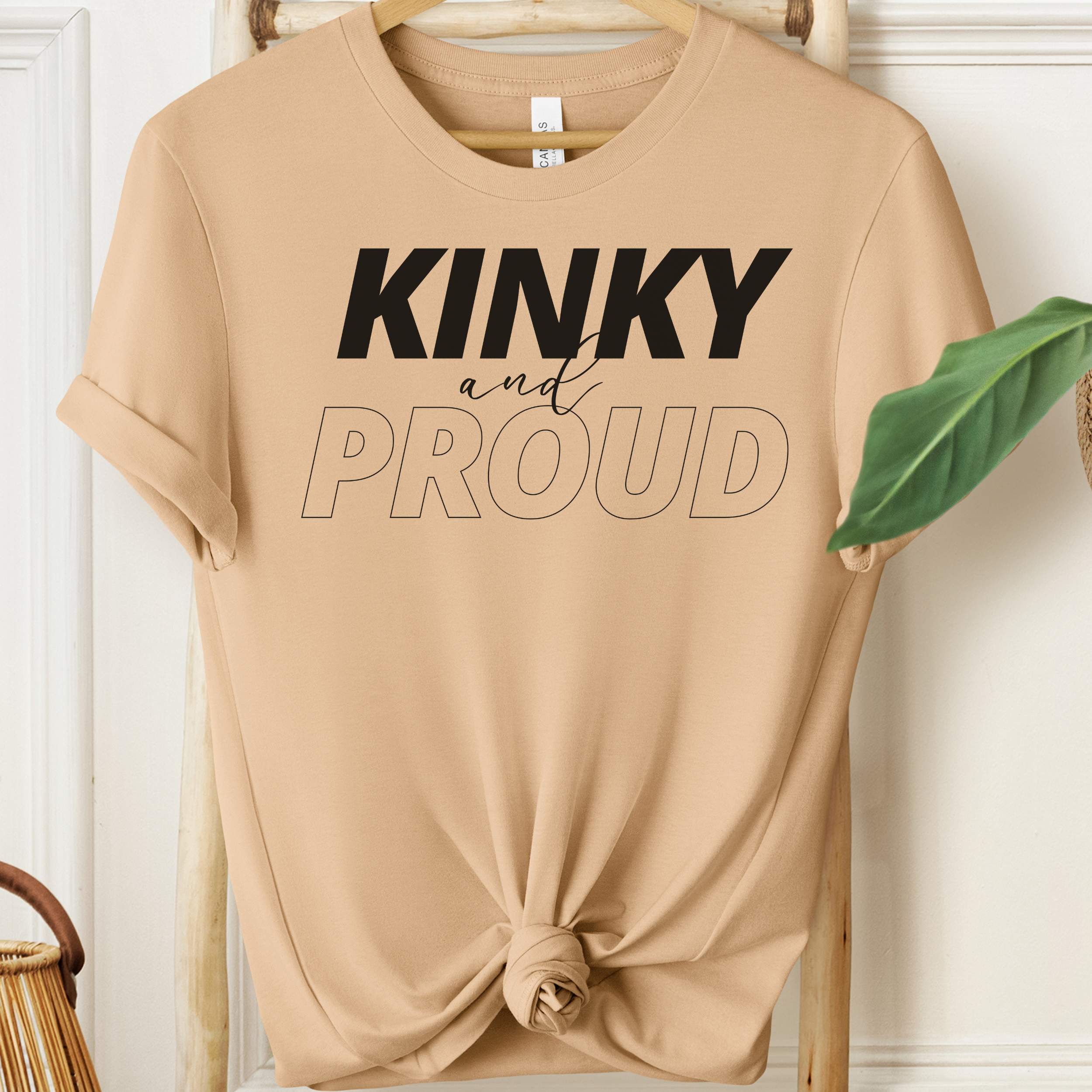 Beige t-shirt with 'Kinky and Proud' text on a wooden hanger.