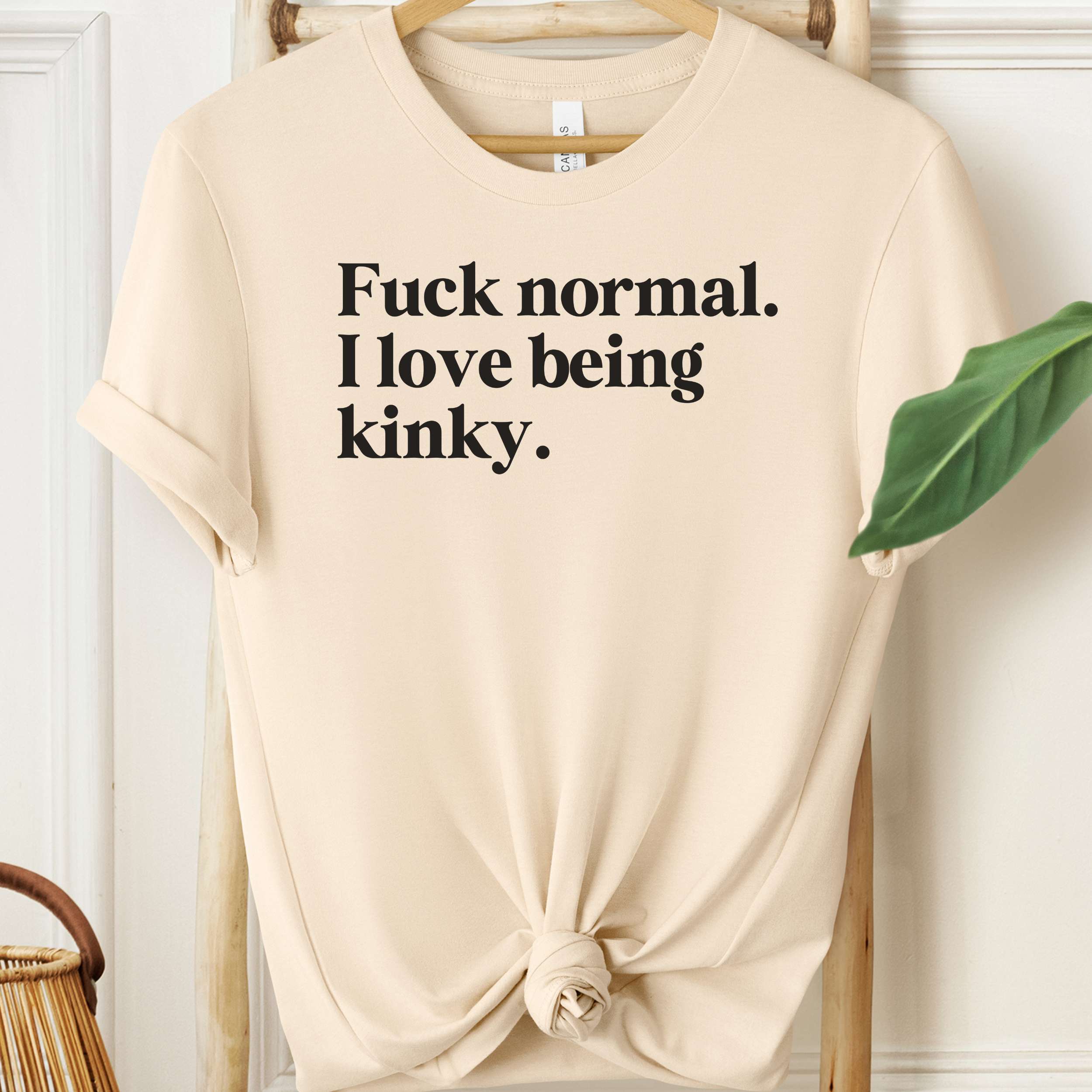 Beige t-shirt with black text on a hanger against a white wall.