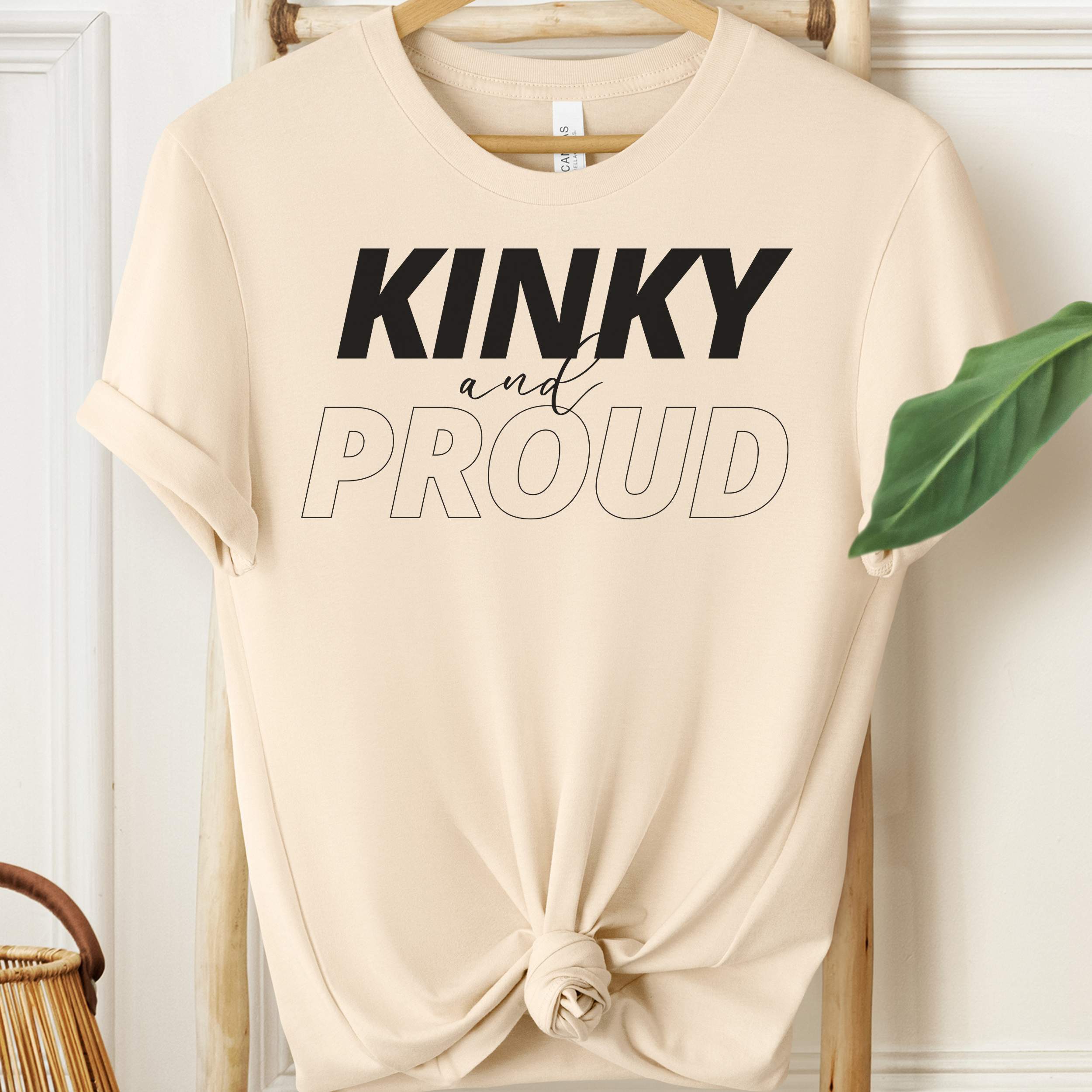Beige t-shirt with 'Kinky and Proud' text on a hanger against a white wall.