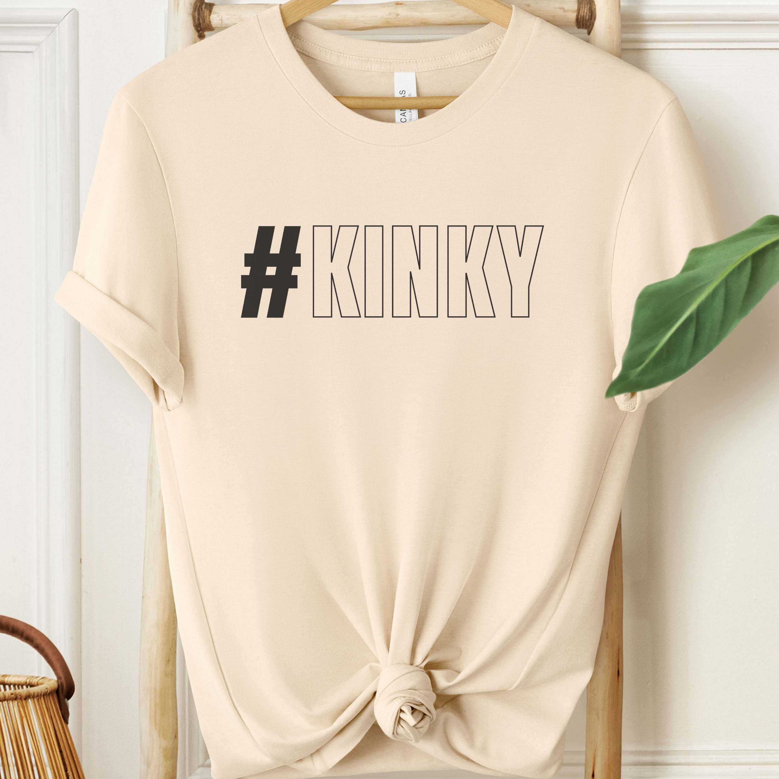Beige t-shirt with '#KINKY' text on a wooden hanger against a white wall.