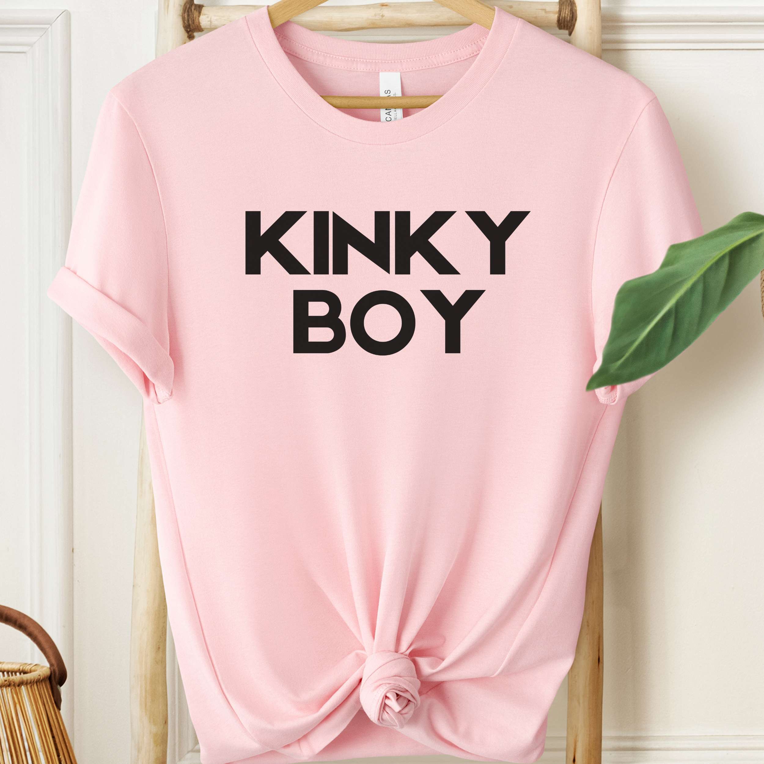 Pink t-shirt with 'KINKY BOY' text on a hanger against a white wall.