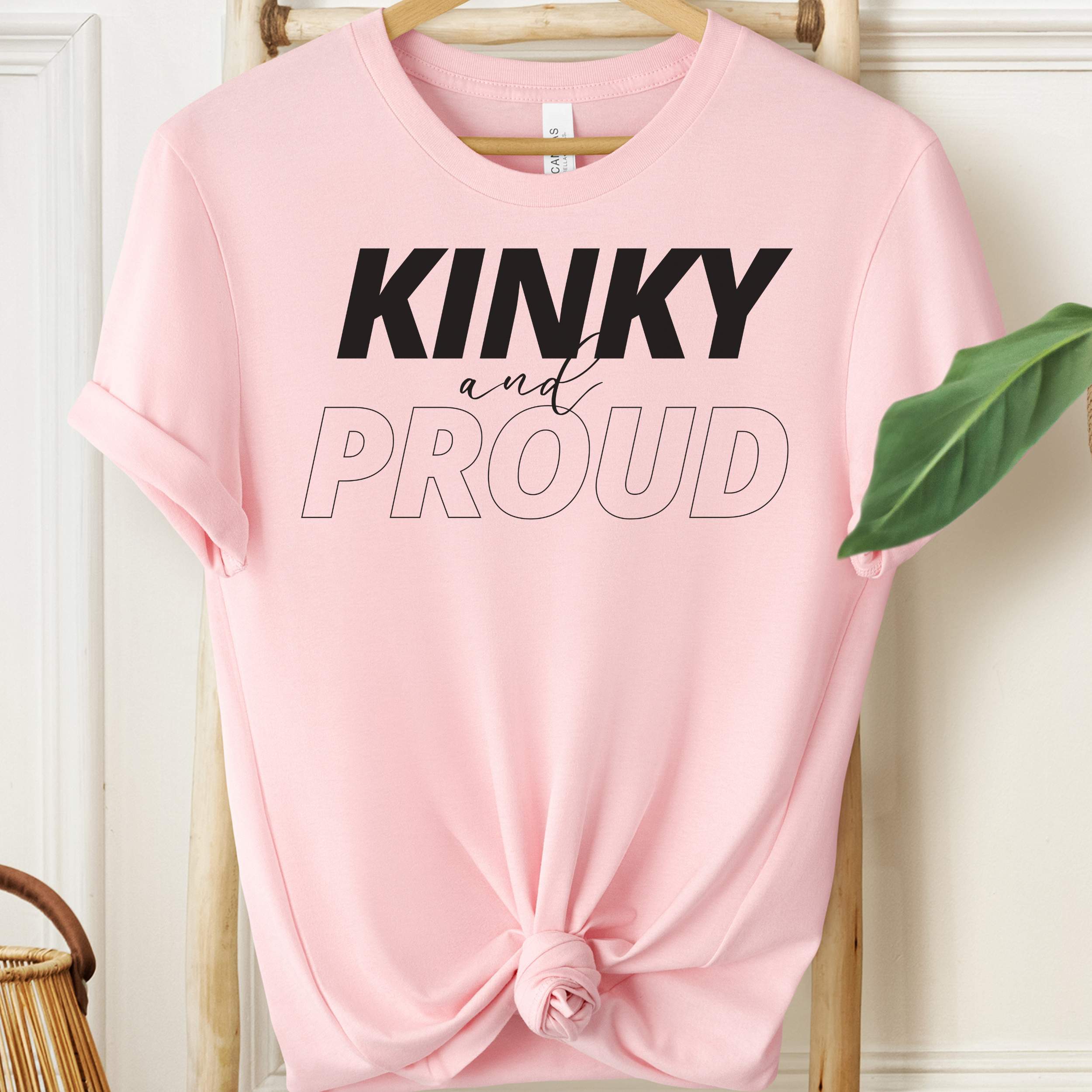 Pink t-shirt with 'Kinky and Proud' text on a hanger against a white wall.