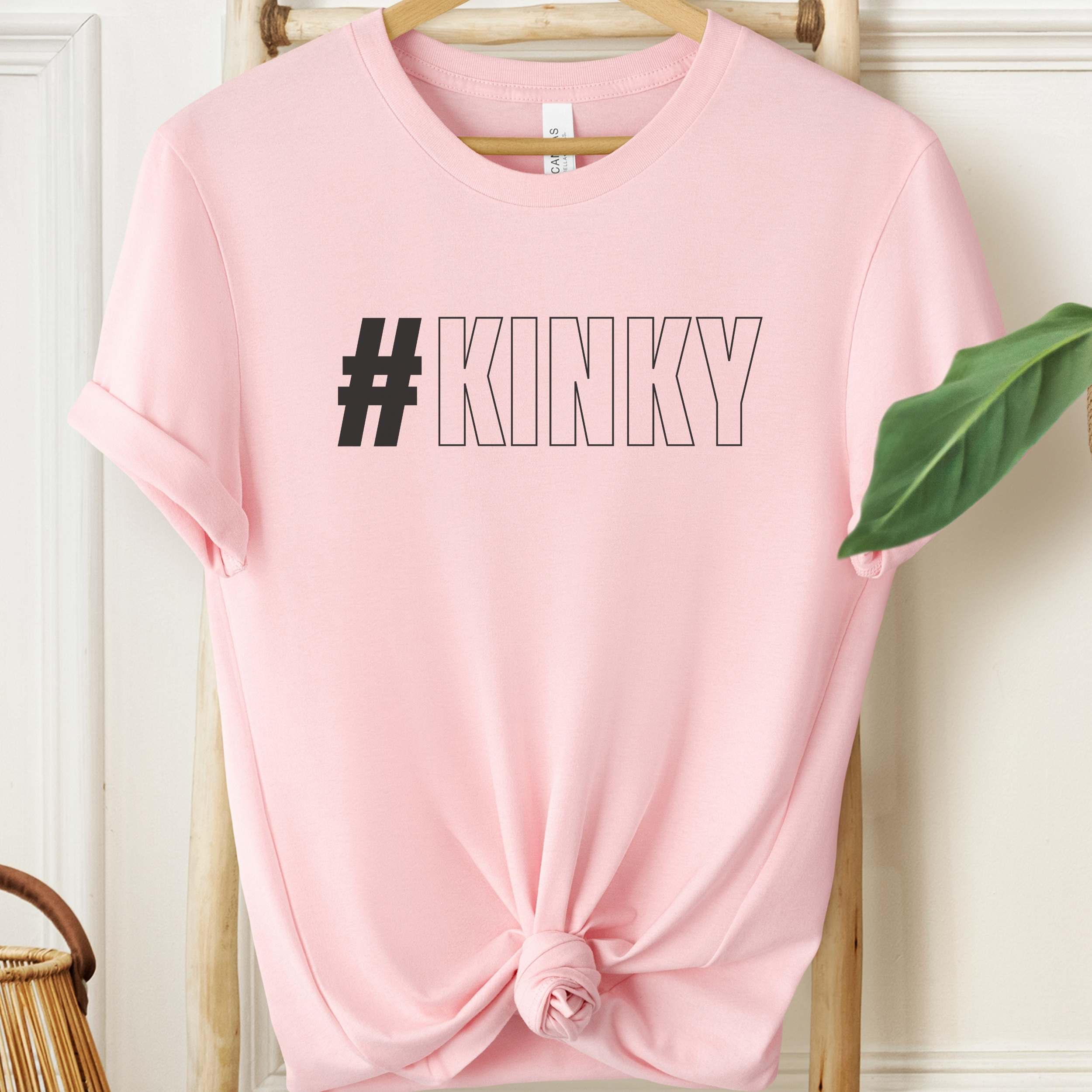 Pink t-shirt with '#KINKY' text on a hanger against a white wall.