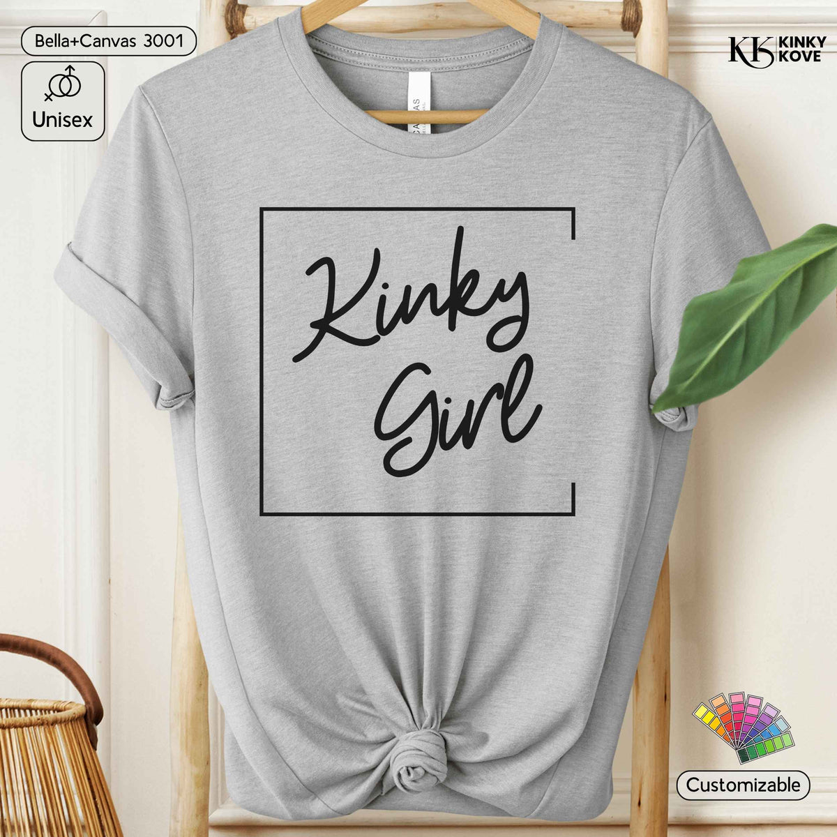 A light heather gray short-sleeve t-shirt with a black graphic print reading 'Kinky Girl' inside a rectangular border, styled on a wooden hanger with a knotted hem. The image includes decorative props like a green leaf and a wicker basket, along with branding overlays for 'Kinky Kove' and 'Bella+Canvas'.