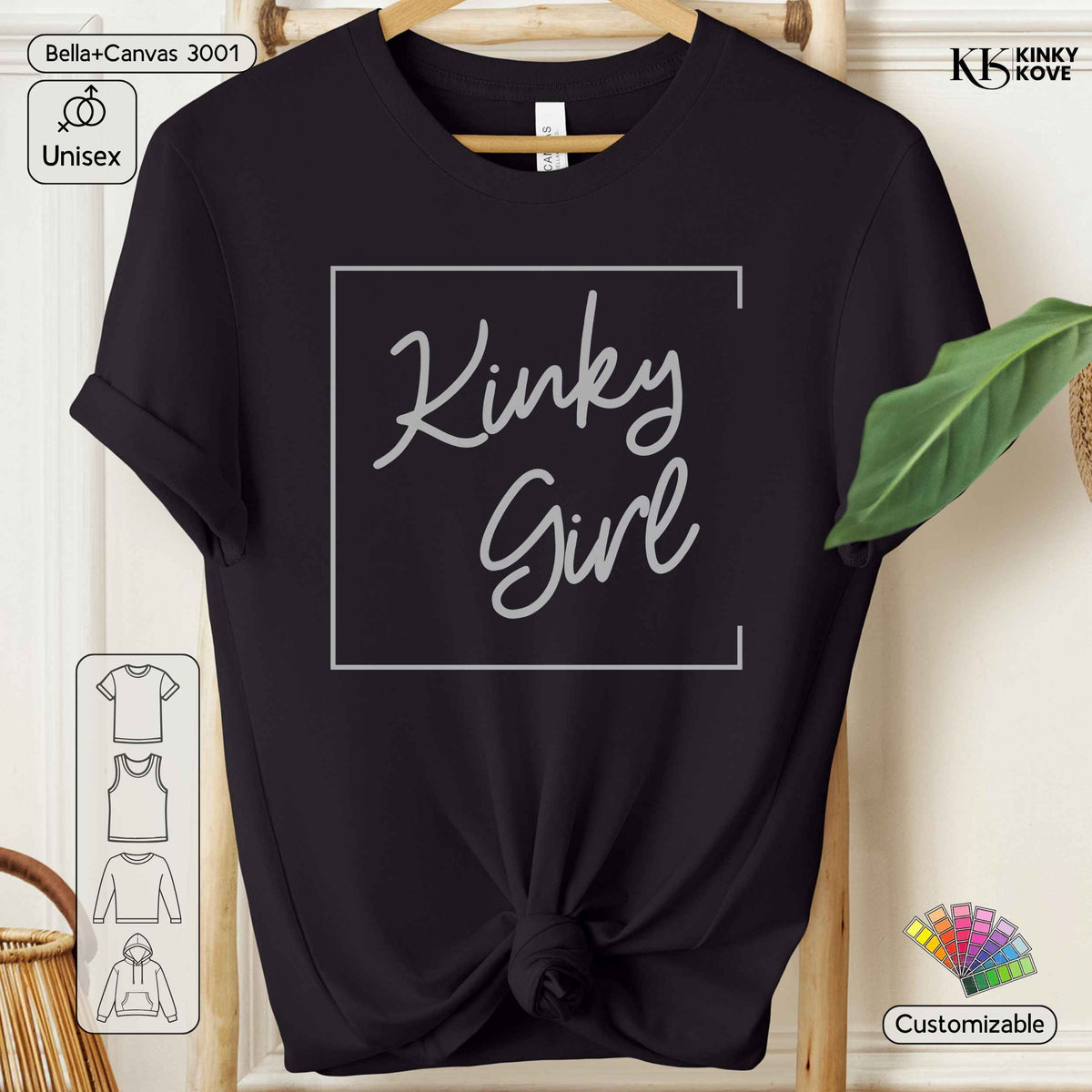 A black short-sleeve t-shirt with a white graphic reading 'Kinky Girl' inside a thin rectangular border, shown hanging on a wooden hanger against a light beige background with decorative props.