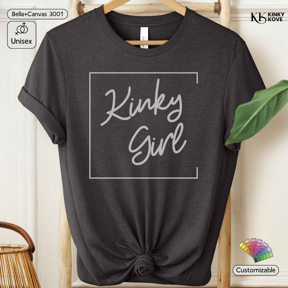A dark heather gray t-shirt with a knotted hem and rolled sleeves, featuring a white graphic that reads 'Kinky Girl' inside a thin square border. The shirt is displayed on a wooden hanger against a neutral background with decorative props.