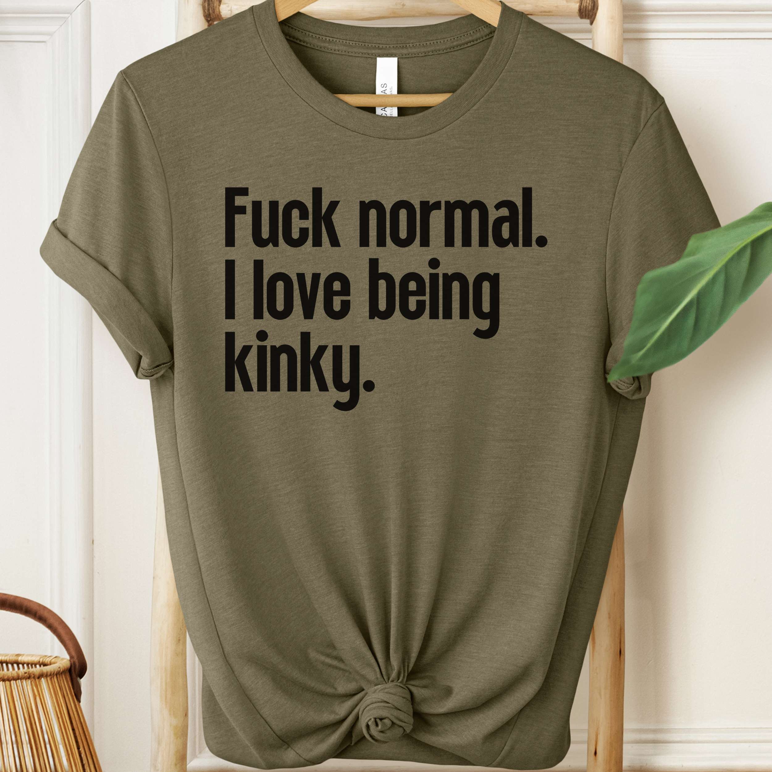 Olive green t-shirt with black text on a hanger against a white wall.