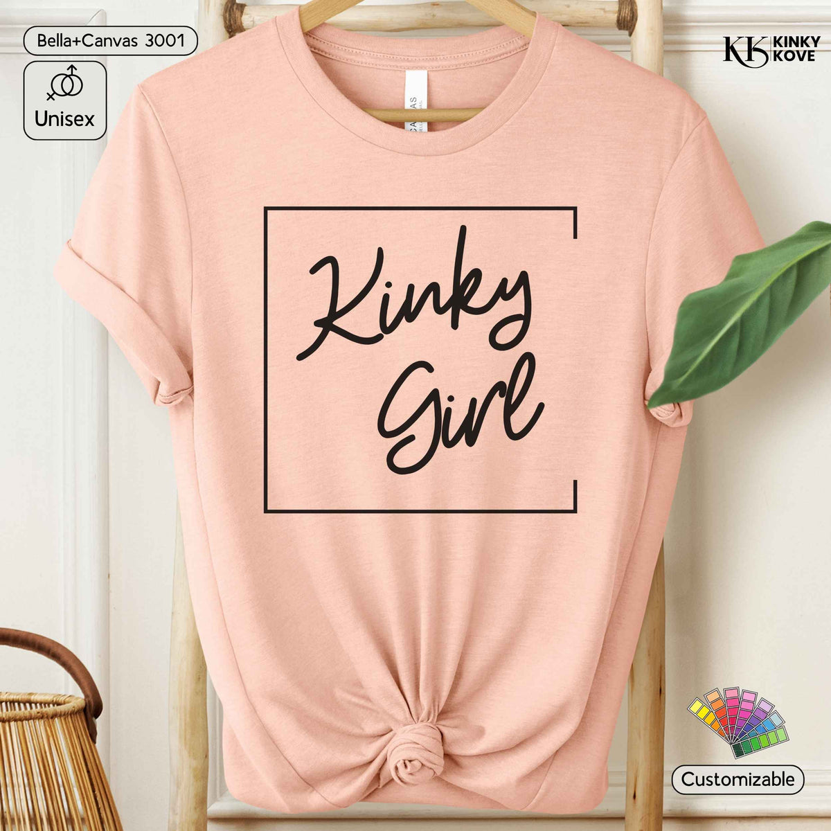 A light pink short-sleeve t-shirt with a black graphic print reading 'Kinky Girl' inside a square border, styled on a wooden hanger with a knotted hem against a neutral indoor backdrop.