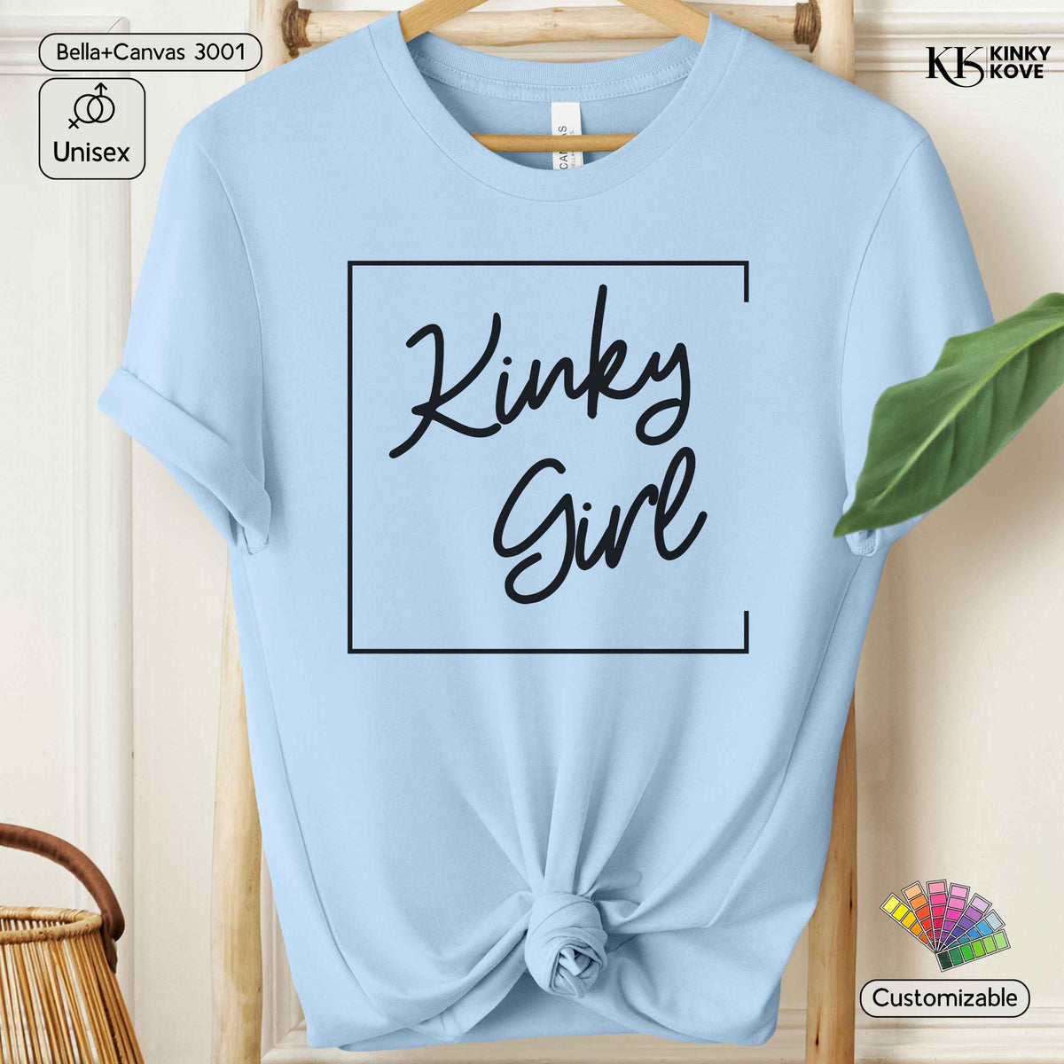 A light blue short-sleeve t-shirt hanging on a wooden hanger, styled with a knot at the bottom hem. The shirt features a black graphic print that reads 'Kinky Girl' inside a thin black square border.