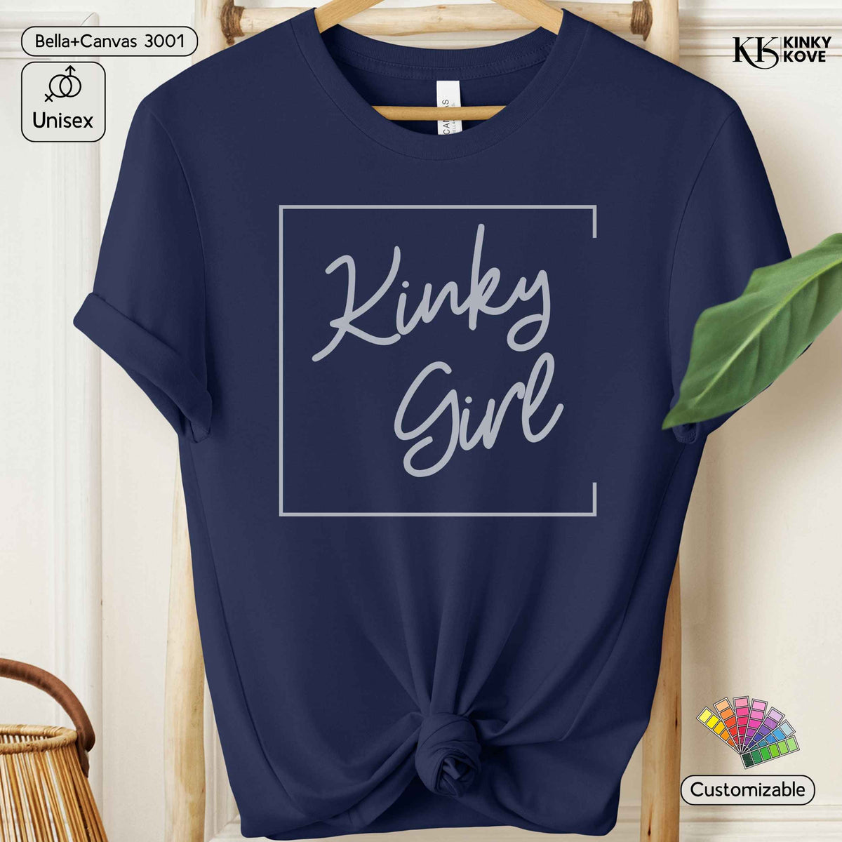 A navy blue short-sleeve t-shirt with a white graphic print reading 'Kinky Girl' inside a thin rectangular border, styled on a wooden hanger with a knotted hem. The background is a warm beige wall with a green leaf and a wicker basket visible.