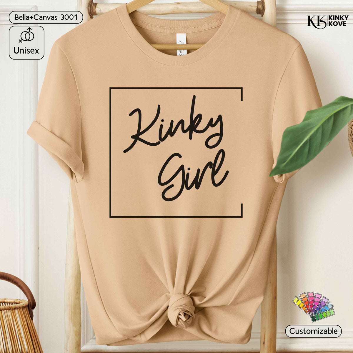A beige short-sleeve t-shirt with rolled cuffs, featuring a black graphic print that reads 'Kinky Girl' inside a square border, styled on a wooden hanger with a knot at the bottom. The background is a white paneled wall with a green leaf prop.