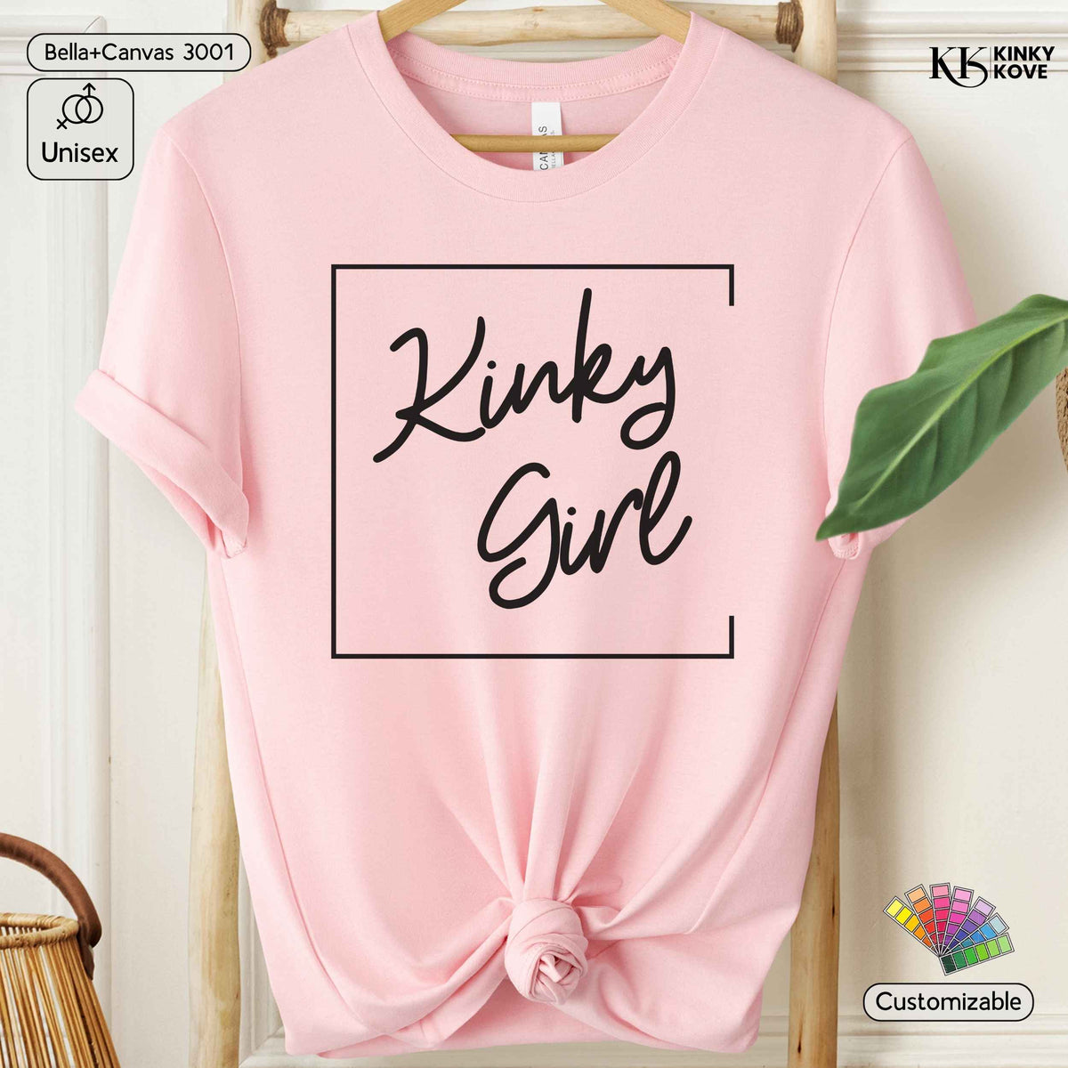 A light pink short-sleeve t-shirt with a knot at the bottom hem, featuring the words 'Kinky Girl' in black script inside a thin black rectangular border on the chest. The t-shirt is displayed on a wooden hanger against a light background with decorative props.