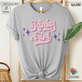 A light heather gray short-sleeve t-shirt with a rolled collar, displayed on a wooden hanger with a knotted hem. The front features a retro-style graphic reading 'Kinky Girl' in pink bubble letters with purple star accents.