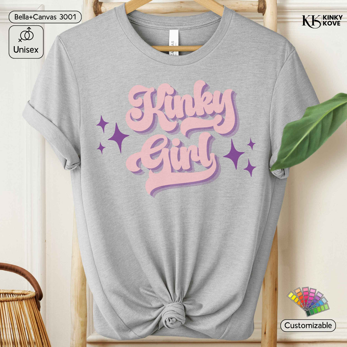 A light heather gray short-sleeve t-shirt with a rolled collar, displayed on a wooden hanger with a knotted hem. The front features a retro-style graphic reading 'Kinky Girl' in pink bubble letters with purple star accents.