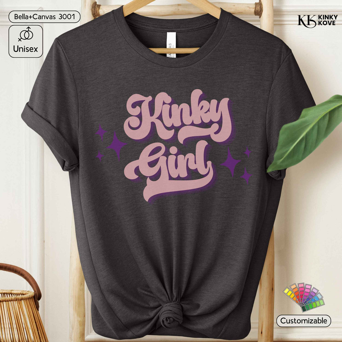 A dark charcoal gray t-shirt with rolled sleeves and a knotted hem, featuring a retro-style pink and purple graphic that reads 'Kinky Girl' with star accents. The shirt is displayed on a wooden hanger against a neutral background with decorative props.