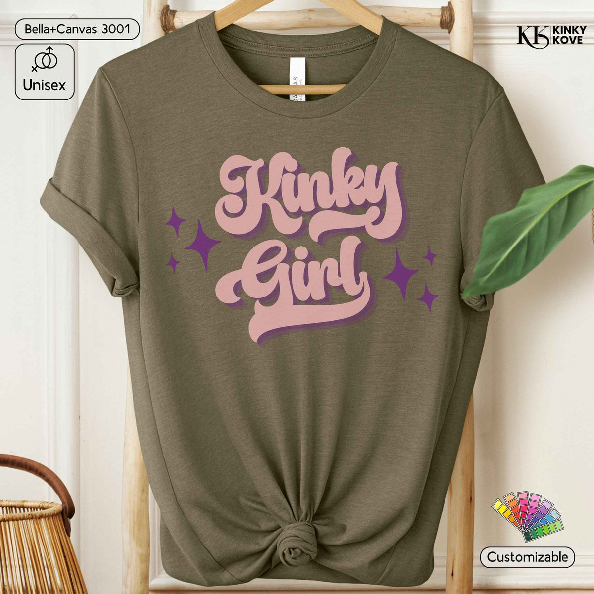 An olive green short-sleeve t-shirt with a knotted hem, featuring a retro-style pink and purple graphic that reads 'Kinky Girl' with star accents. The t-shirt is displayed on a wooden hanger rack with a green leaf prop, set against a light wood panel background.