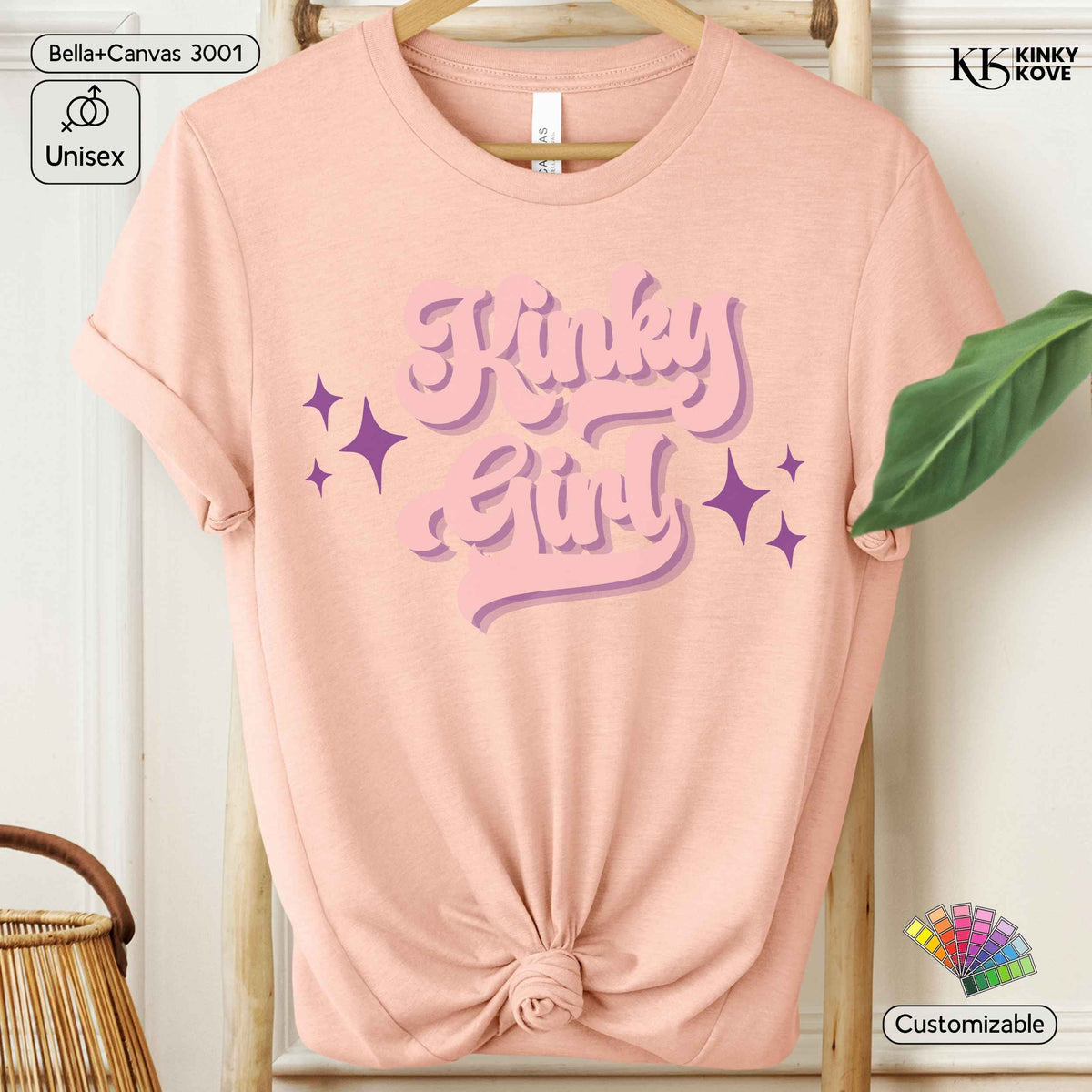 A light peach-pink short-sleeve t-shirt with a knotted hem, featuring a retro-style purple graphic that reads 'Kinky Girl' with purple sparkles. The shirt is displayed on a wooden hanger against a white paneled wall, with decorative props including a green leaf, a woven basket, and digital customization badges.