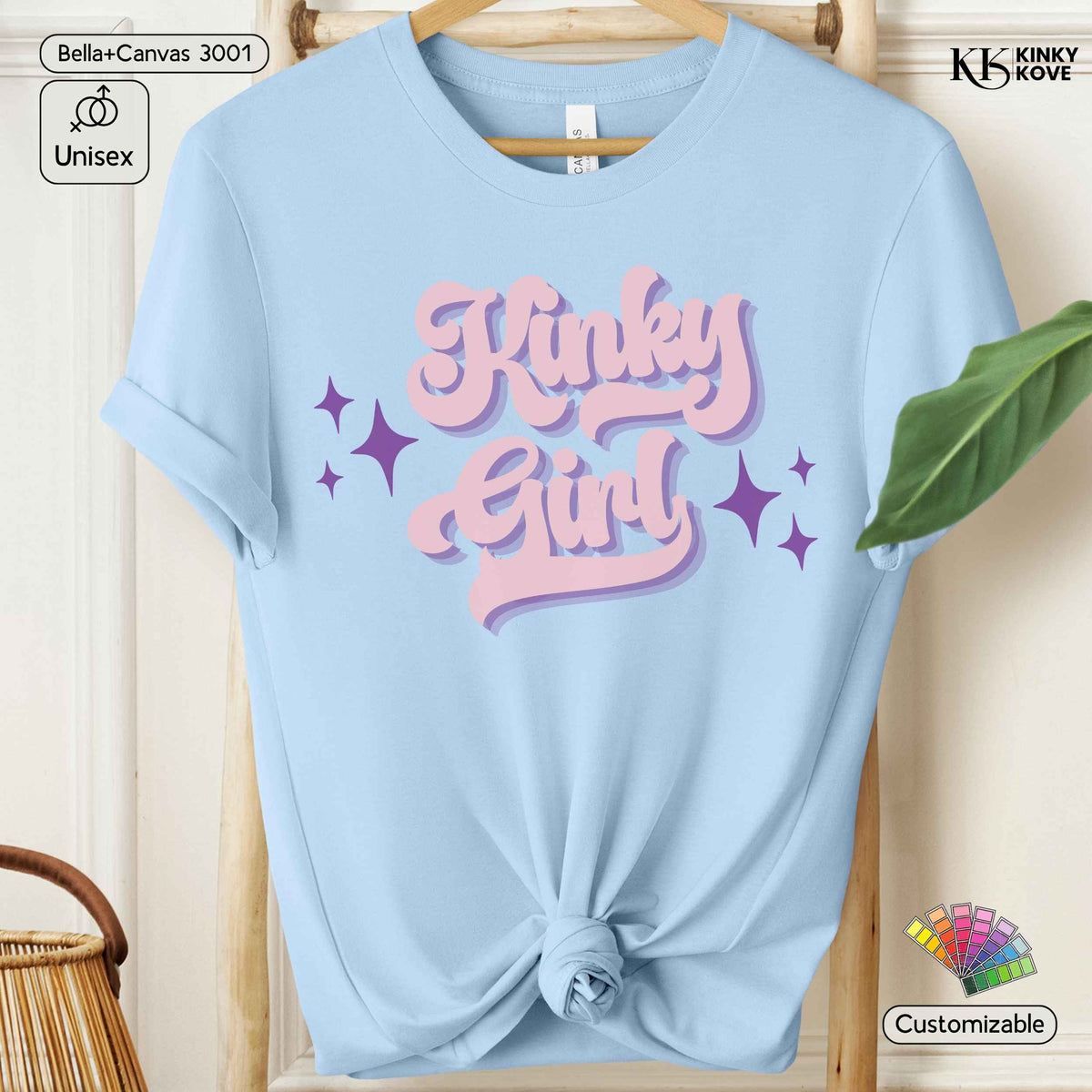A light blue short-sleeve t-shirt with a graphic reading 'Kinky Girl' in pink bubble letters with a purple shadow effect, displayed on a wooden hanger with a knotted hem.