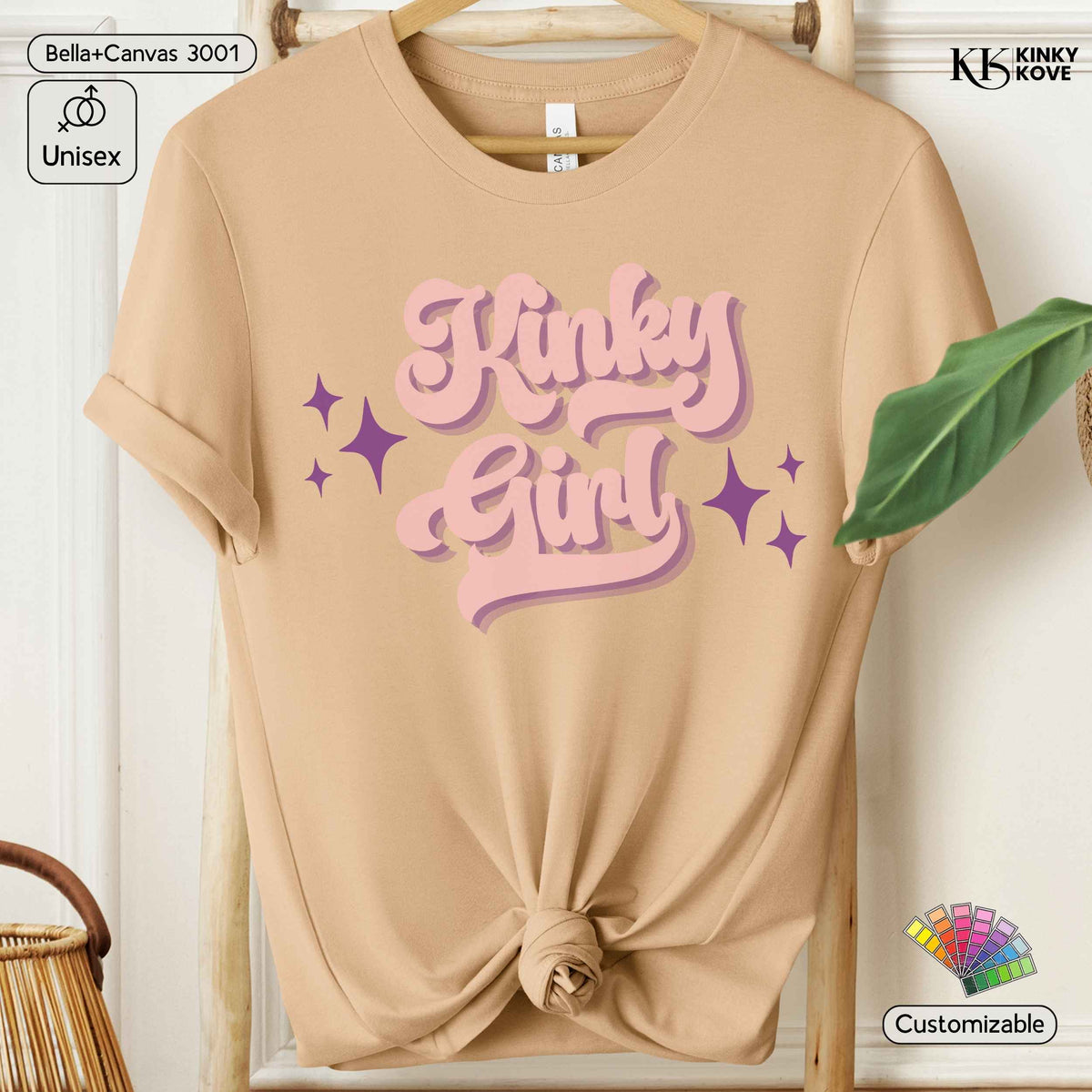 A beige short-sleeve t-shirt hanging on a wooden hanger, styled with a knot at the bottom hem. The shirt features a retro-style graphic on the chest that reads 'Kinky Girl' in pink bubble letters with purple star accents.