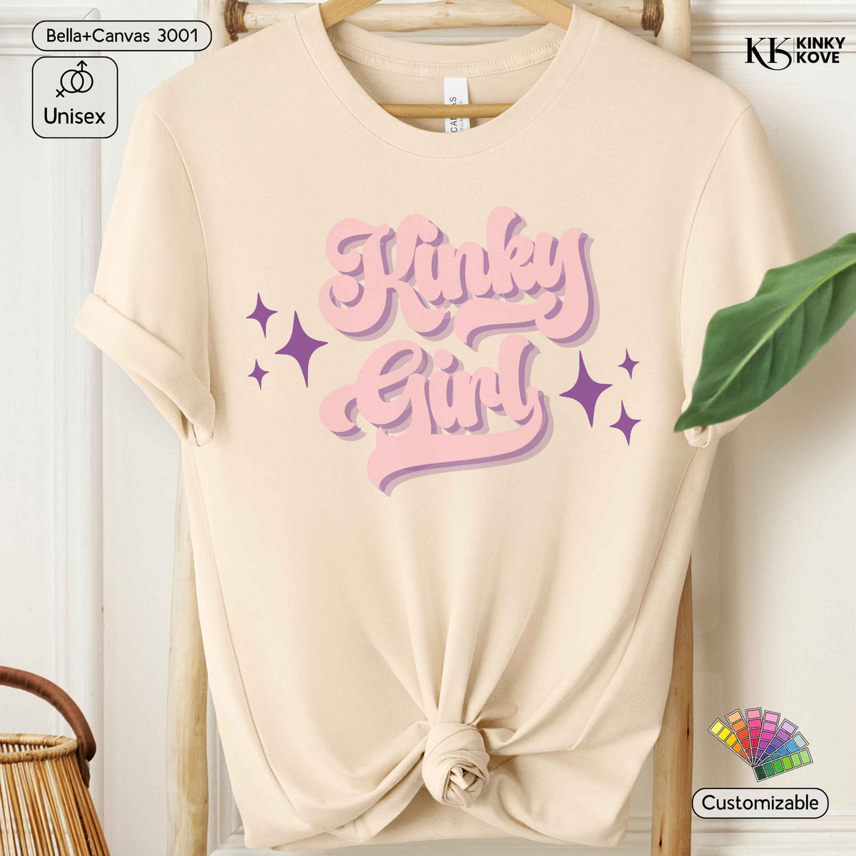 A cream-colored short-sleeve t-shirt with a knotted hem, featuring a pink and purple graphic that reads 'Kinky Girl' with star accents. The t-shirt is displayed on a wooden hanger against a light background with decorative props.