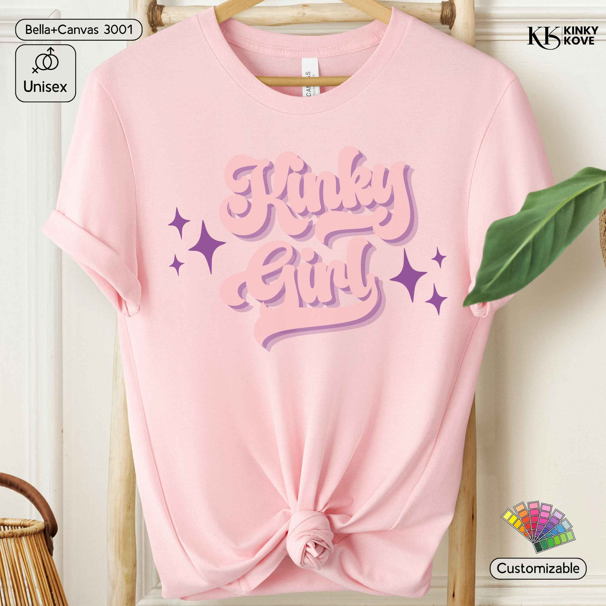 A light pink short-sleeve t-shirt with 'Kinky Girl' printed in a stylized purple and pink font with star accents, hanging on a wooden hanger. The bottom of the shirt is tied in a knot.
