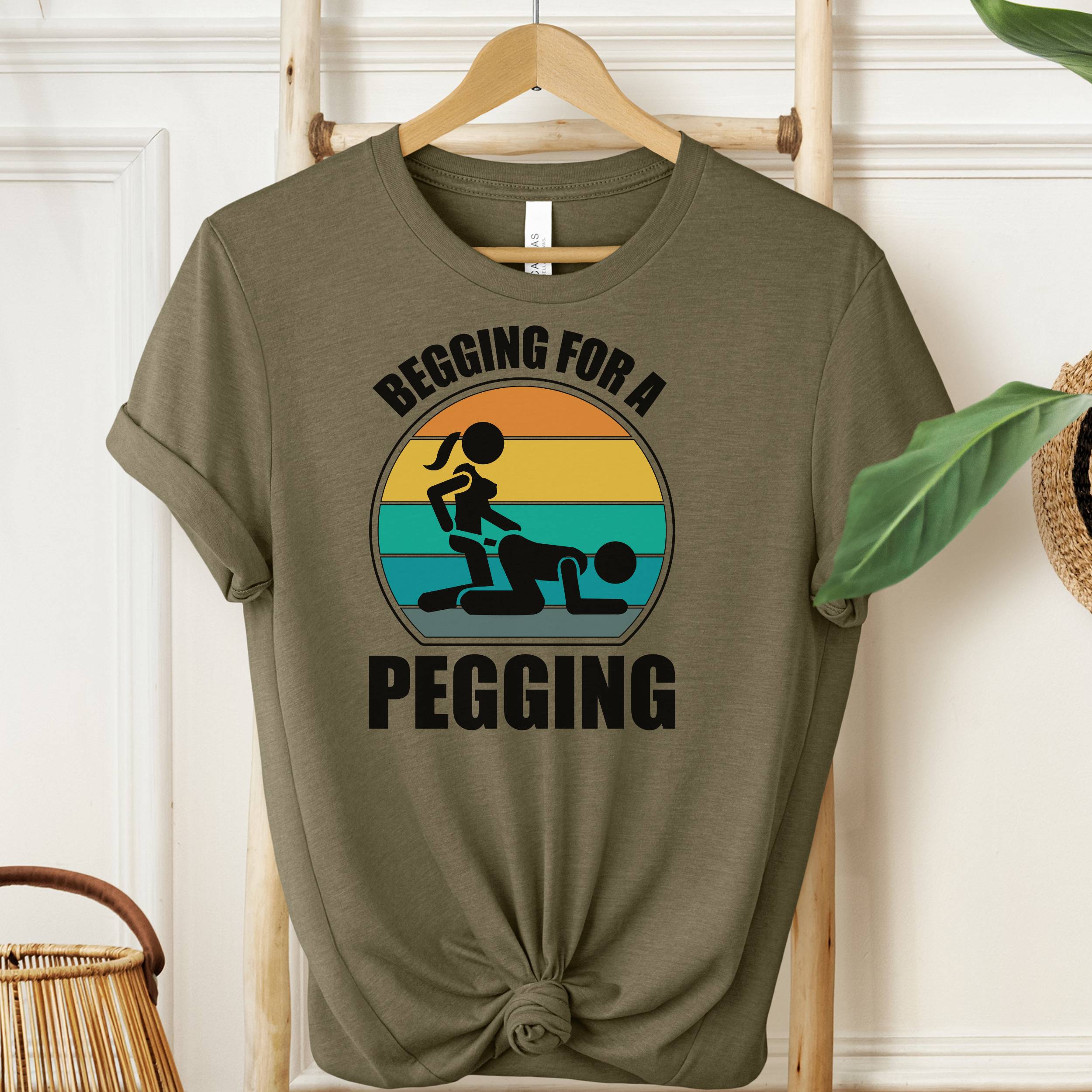 Green t-shirt with graphic design and text 'Begging for a Pegging' on a wooden hanger.