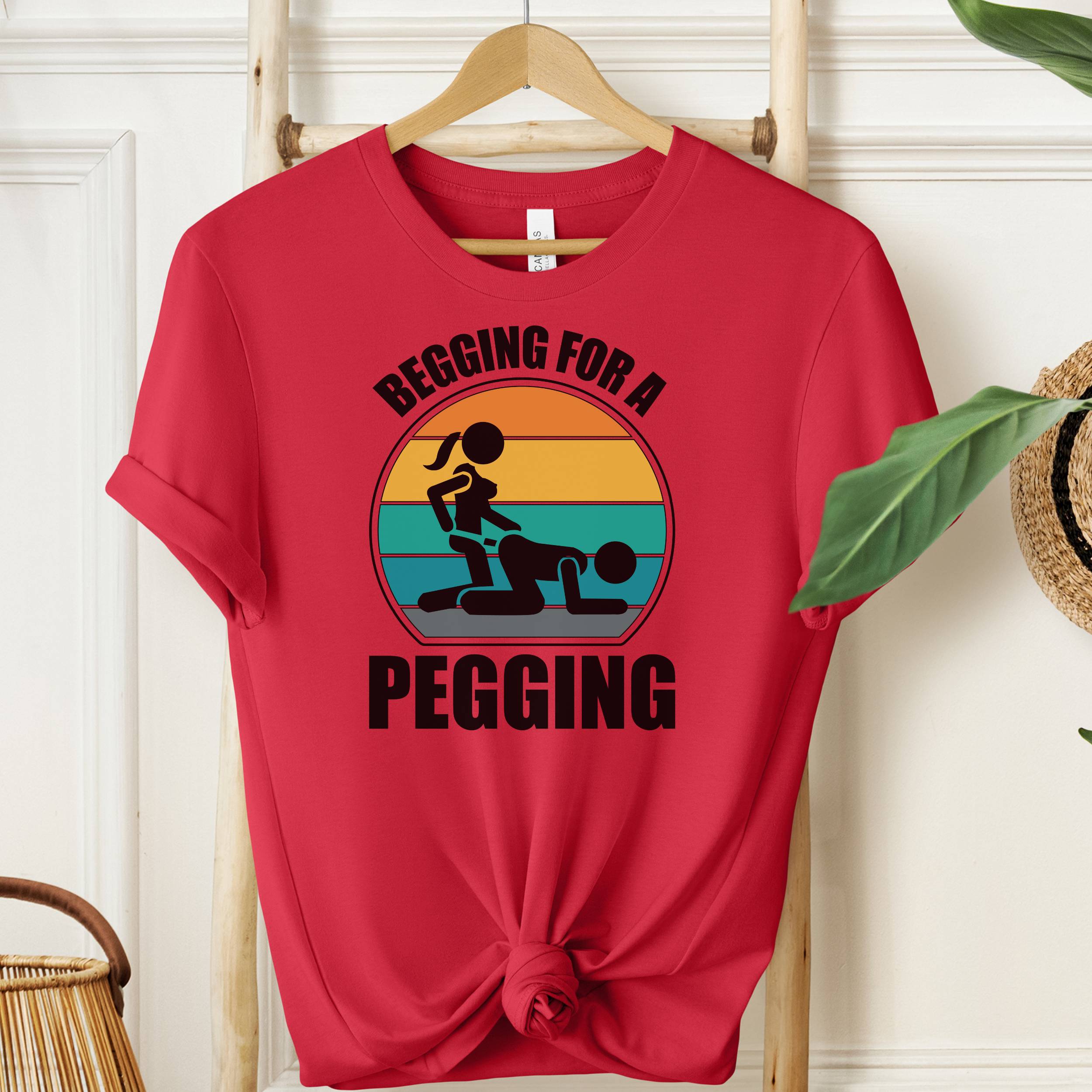 Red t-shirt with graphic design and text 'Begging for a Pegging' on a wooden hanger.