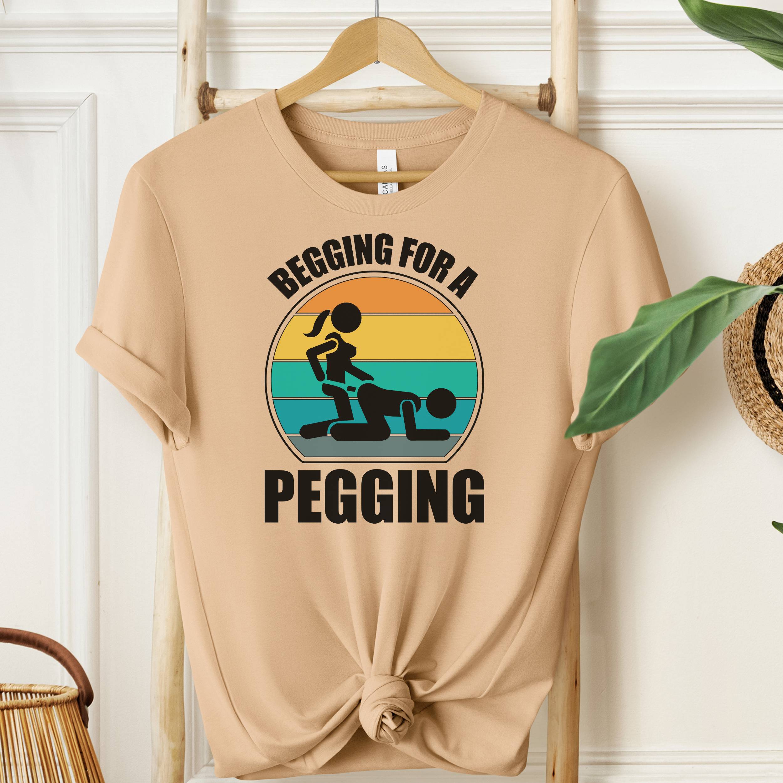 Beige t-shirt with graphic design and text 'Begging for a Pegging' on a wooden hanger.