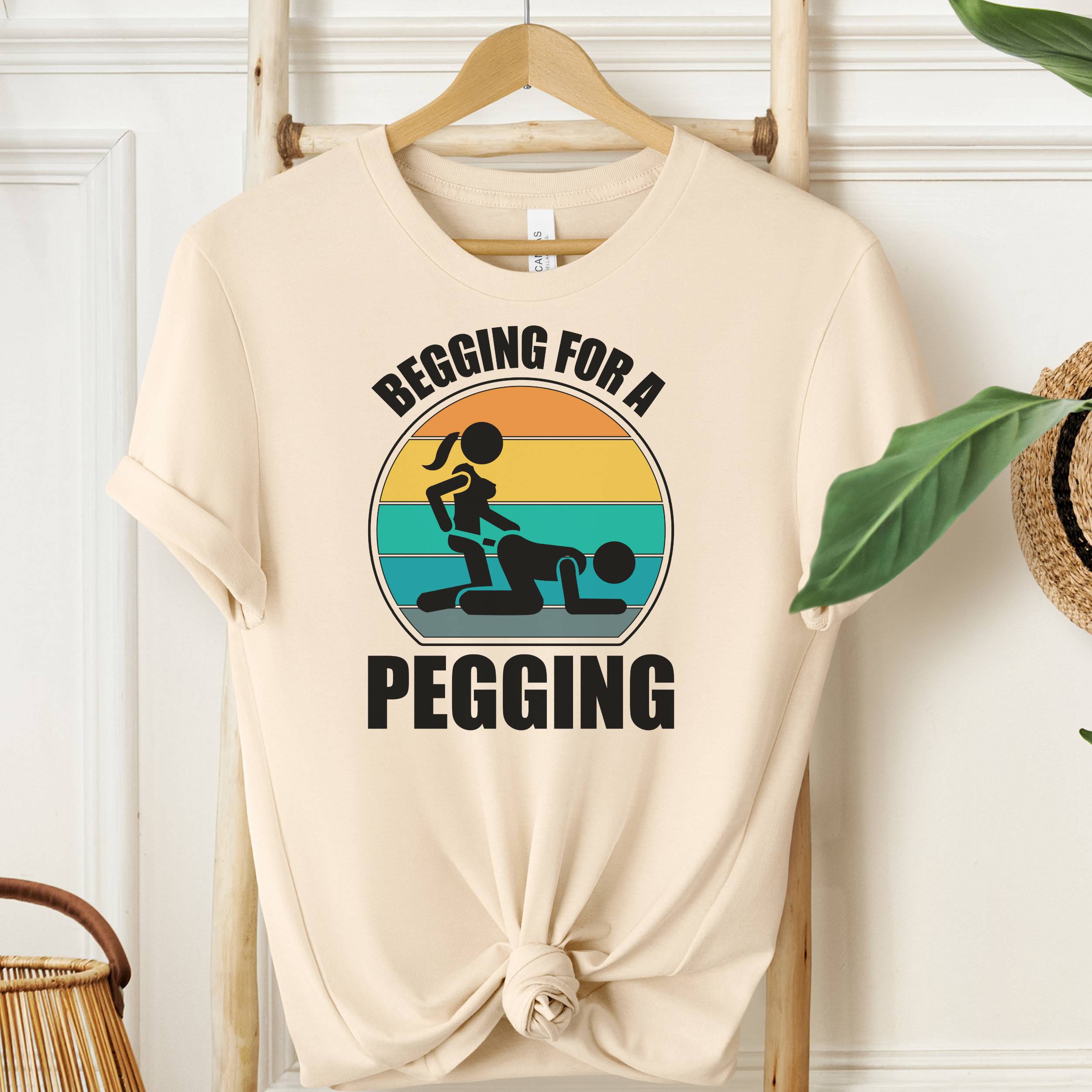 Beige t-shirt with graphic design and text 'Begging for a Pegging' on a wooden hanger.