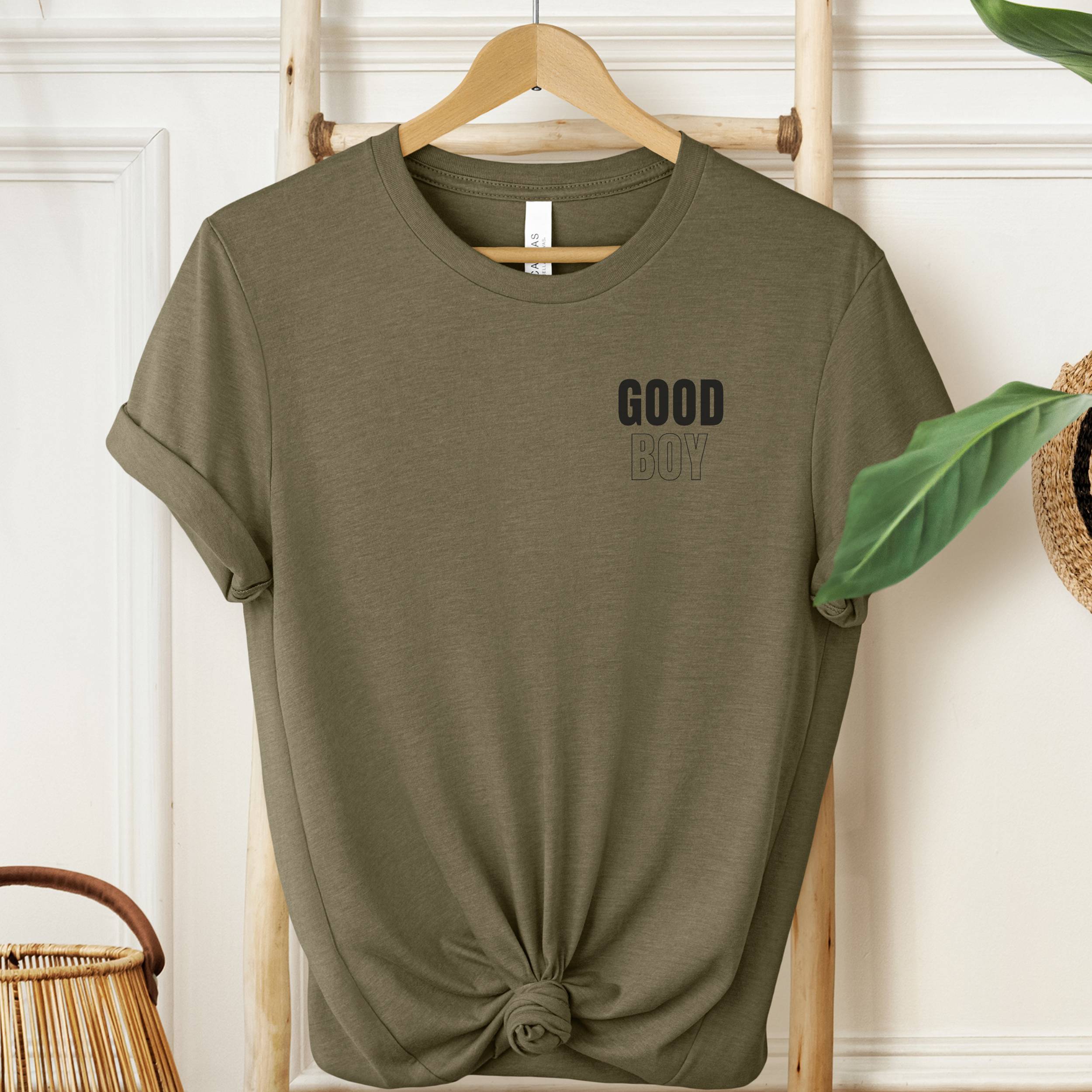Olive green t-shirt with 'GOOD BOY' text on a hanger against a white wall.