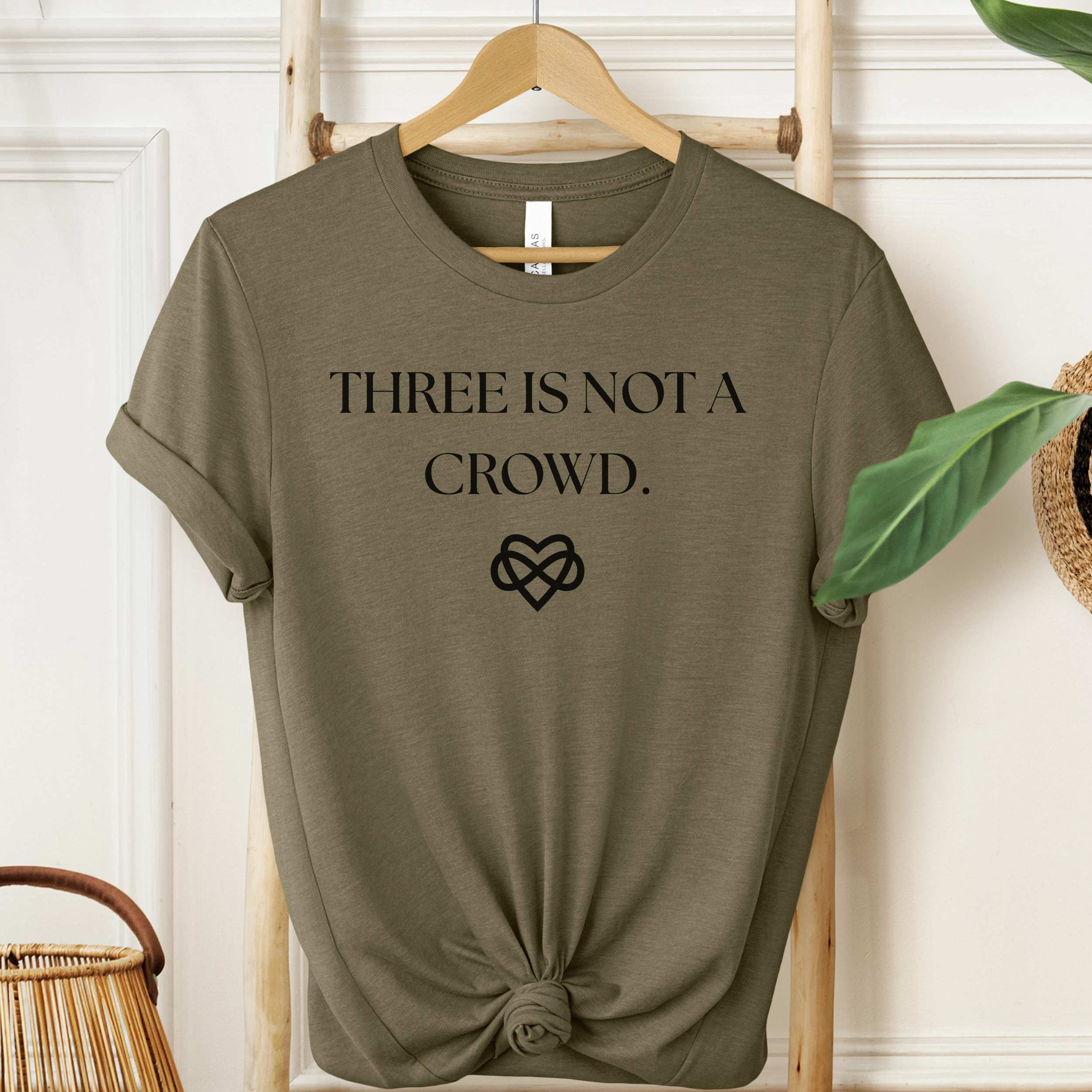 Green t-shirt with 'THREE IS NOT A CROWD' text on a hanger against a white wall.