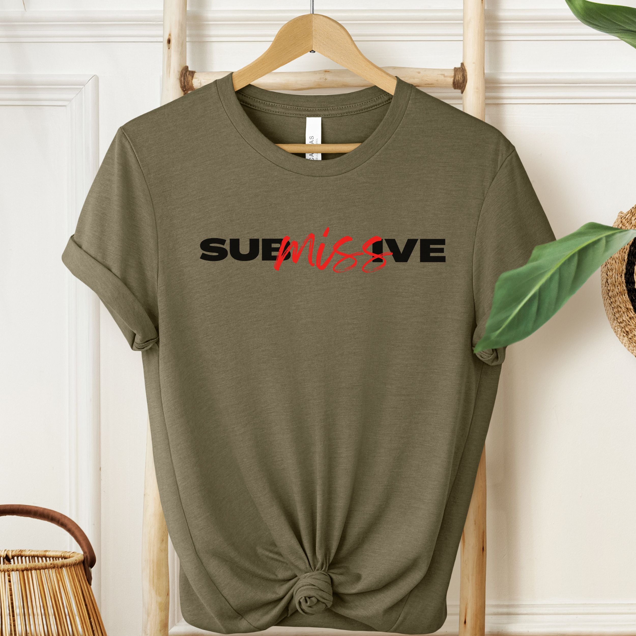 Olive green t-shirt with 'SUBMISSIVE' text on a hanger against a white wall.