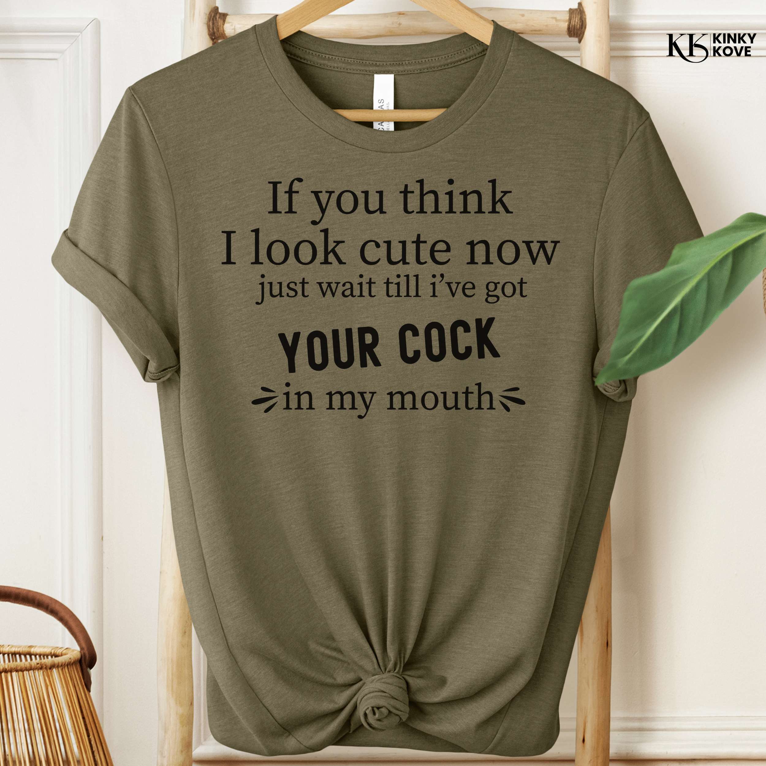 Olive green t-shirt with humorous text on a hanger against a white wall.
