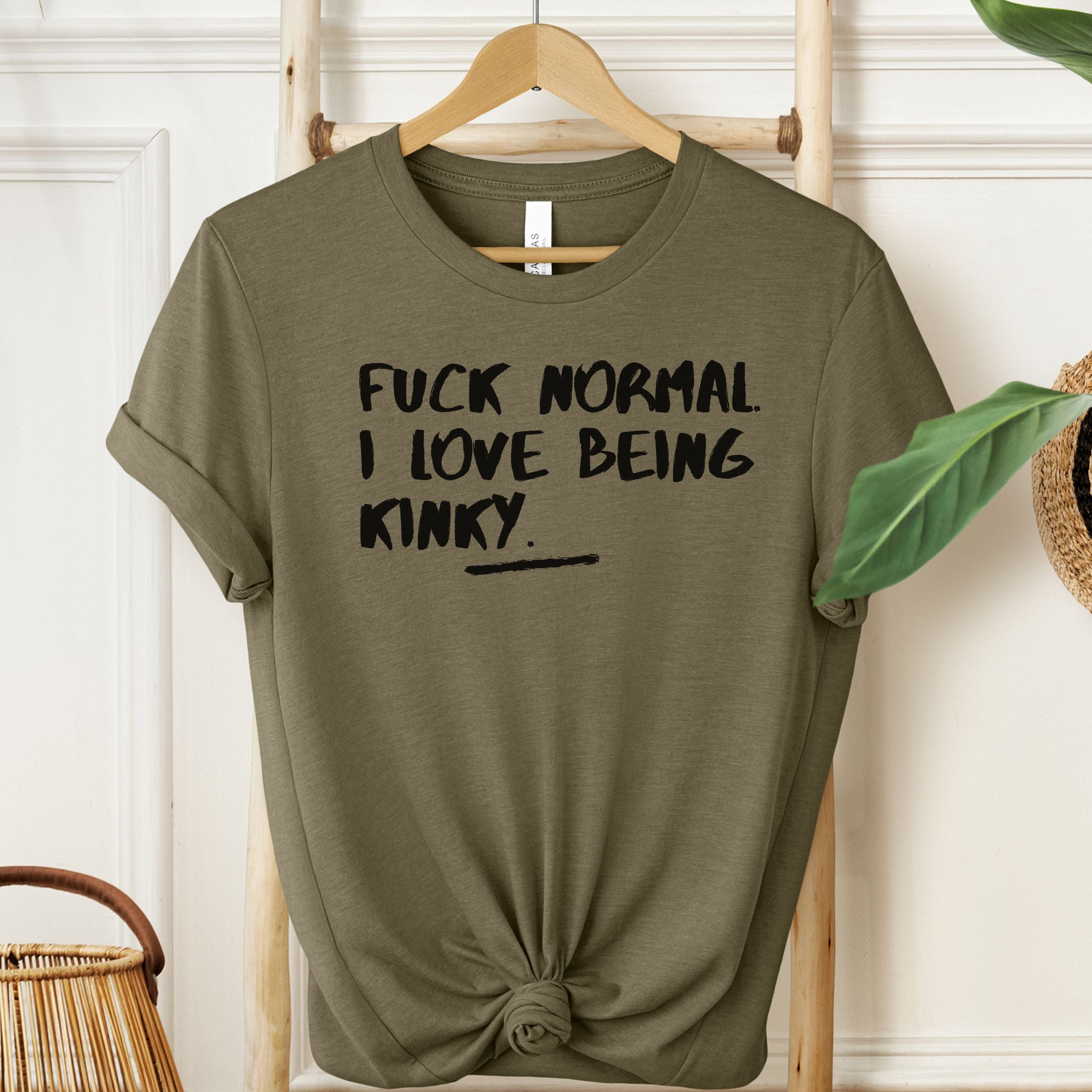 Green t-shirt with text 'FUCK NORMAL I LOVE BEING KINKY' on a hanger against a white wall.