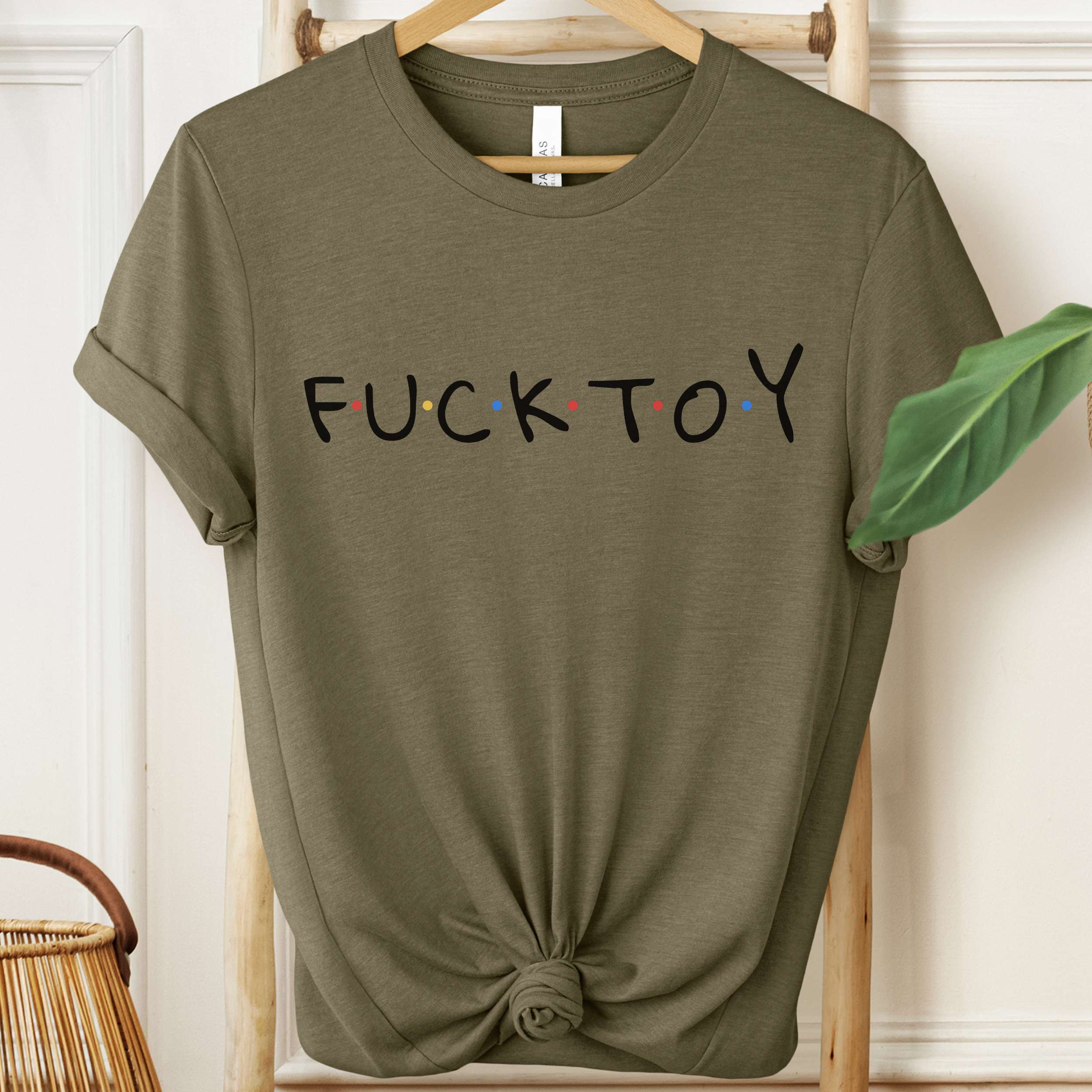 Green t-shirt with 'FUCK TOY' text on a hanger against a white wall.