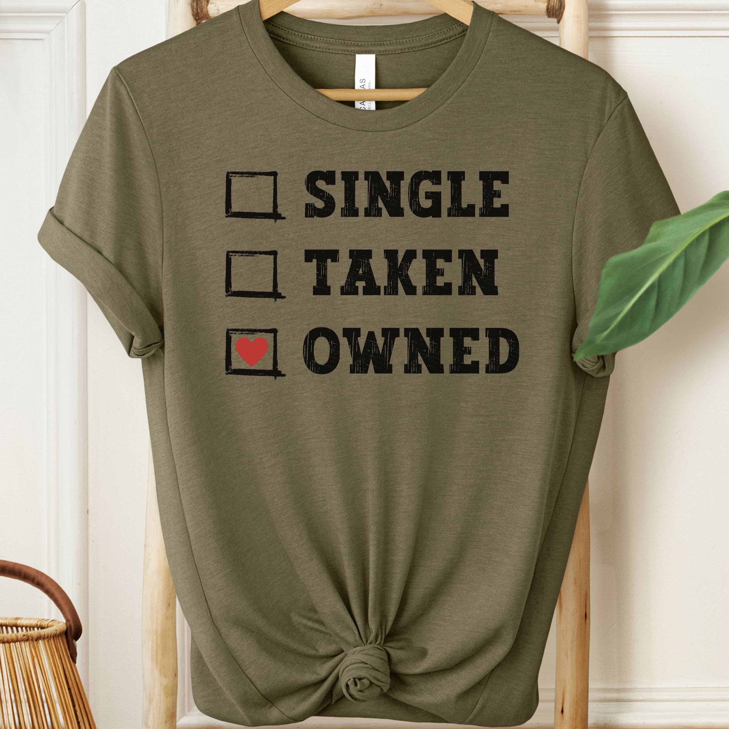 Olive green t-shirt with text 'Single, Taken, Owned' on a hanger.