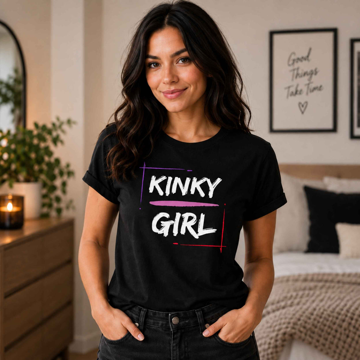 A woman wearing a black graphic t-shirt with 'KINKY GIRL' printed in white text with purple and red accents, standing in a cozy bedroom setting.