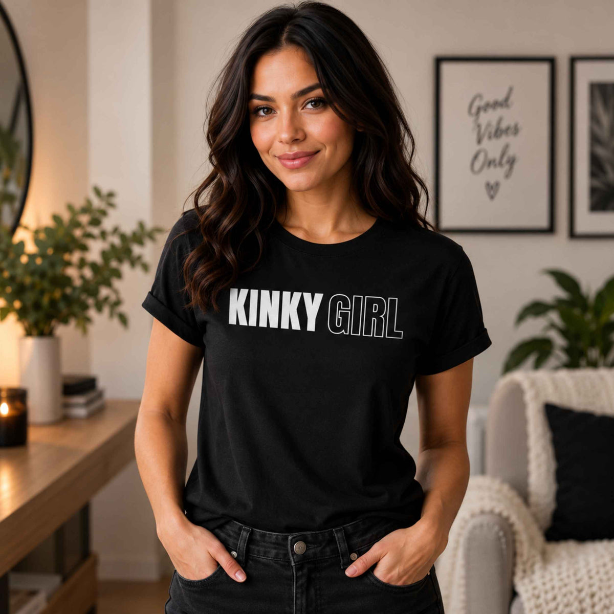 A woman wearing a black short-sleeve t-shirt with the words 'KINKY GIRL' printed across the chest in white and gray, standing in a cozy, modern living room setting.