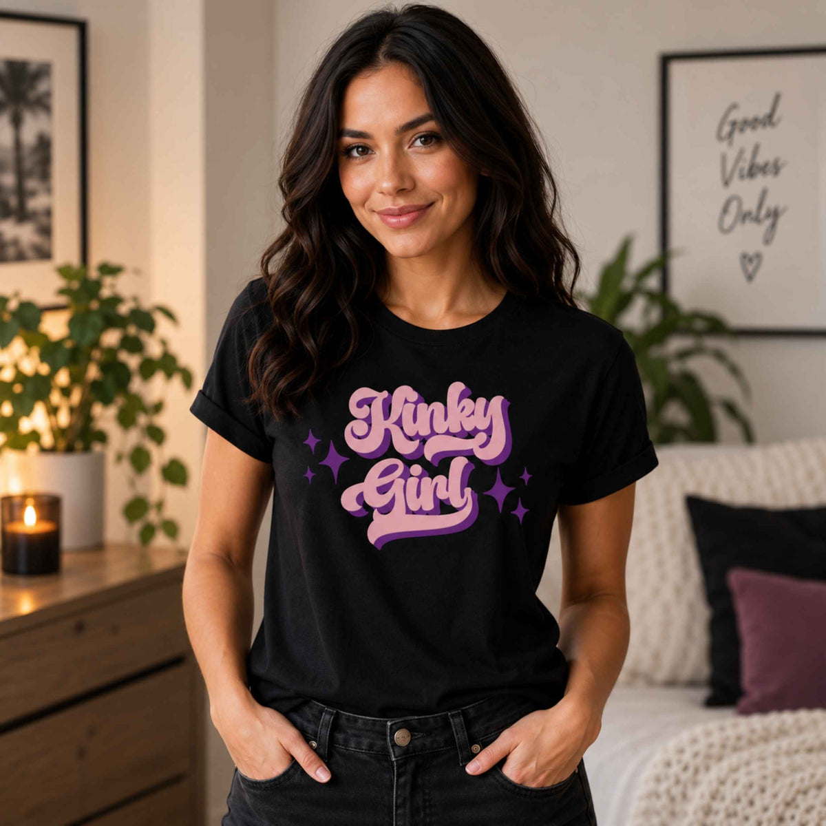 A woman wearing a black graphic t-shirt with 'Rinky Dinky Girl' printed in pink and purple retro-style lettering, standing in a cozy, warmly lit bedroom setting.
