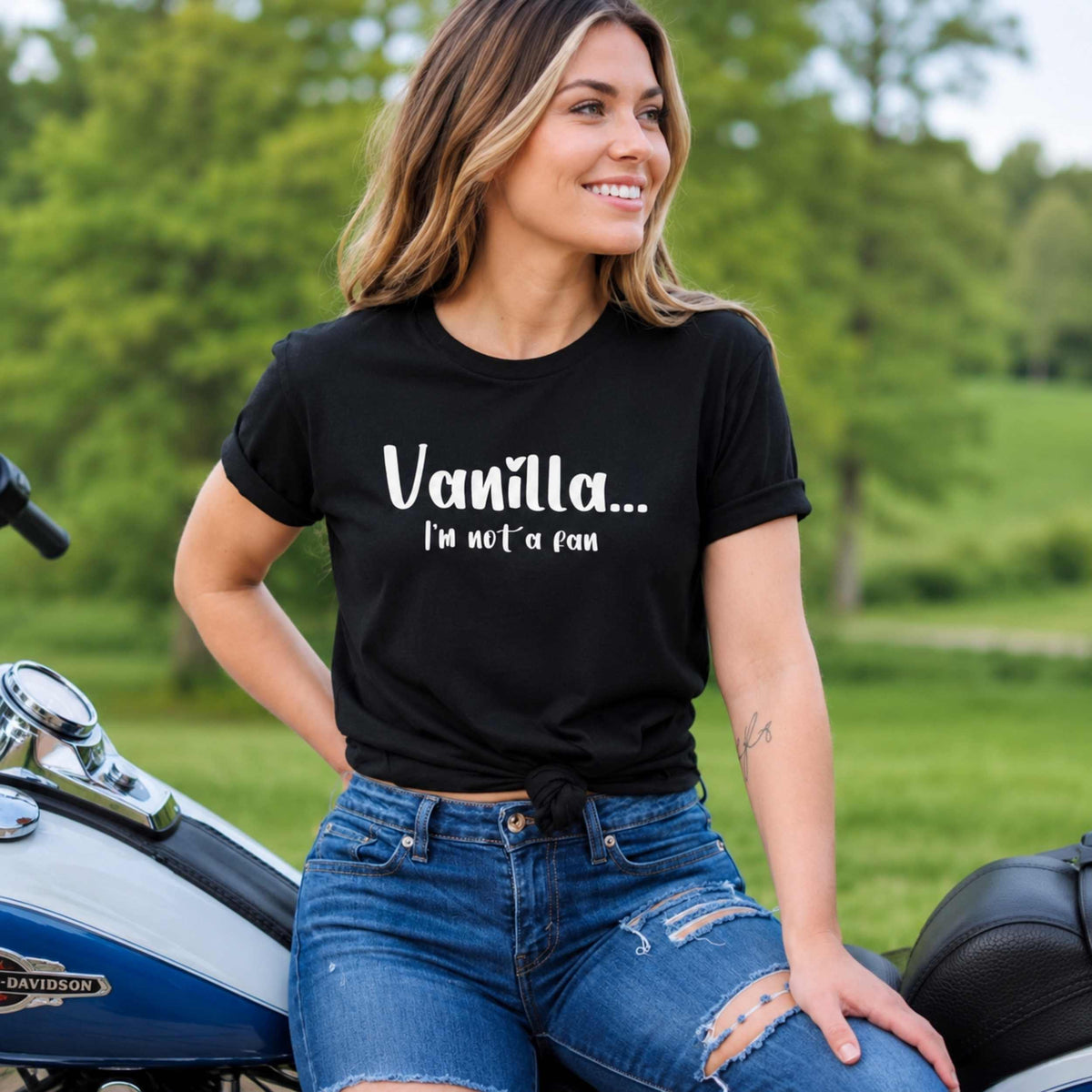 A woman sitting on a motorcycle outdoors wearing a black graphic t-shirt with the text 'Vanilla.
