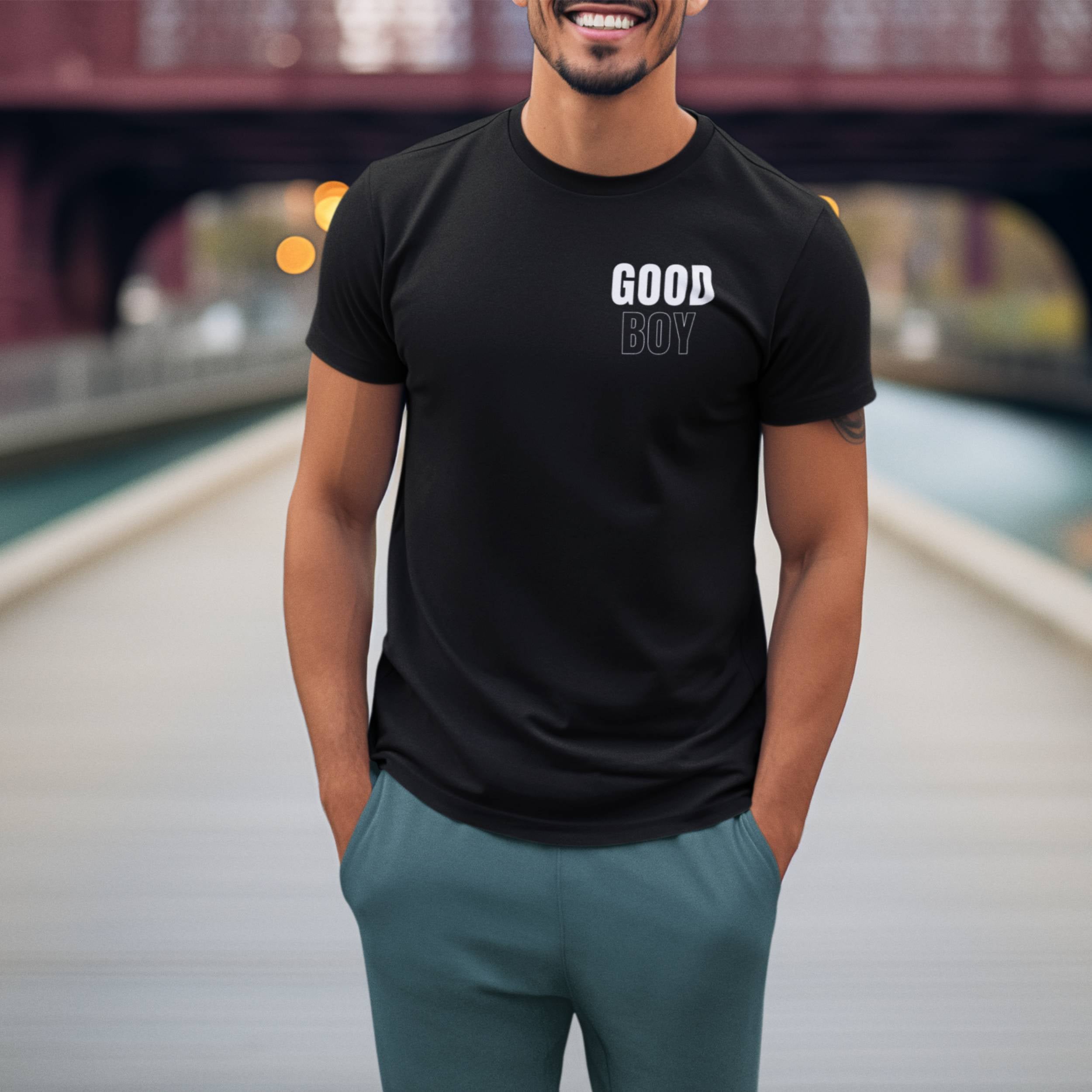 Man wearing a black t-shirt with 'GOOD BOY' text, standing on a bridge.