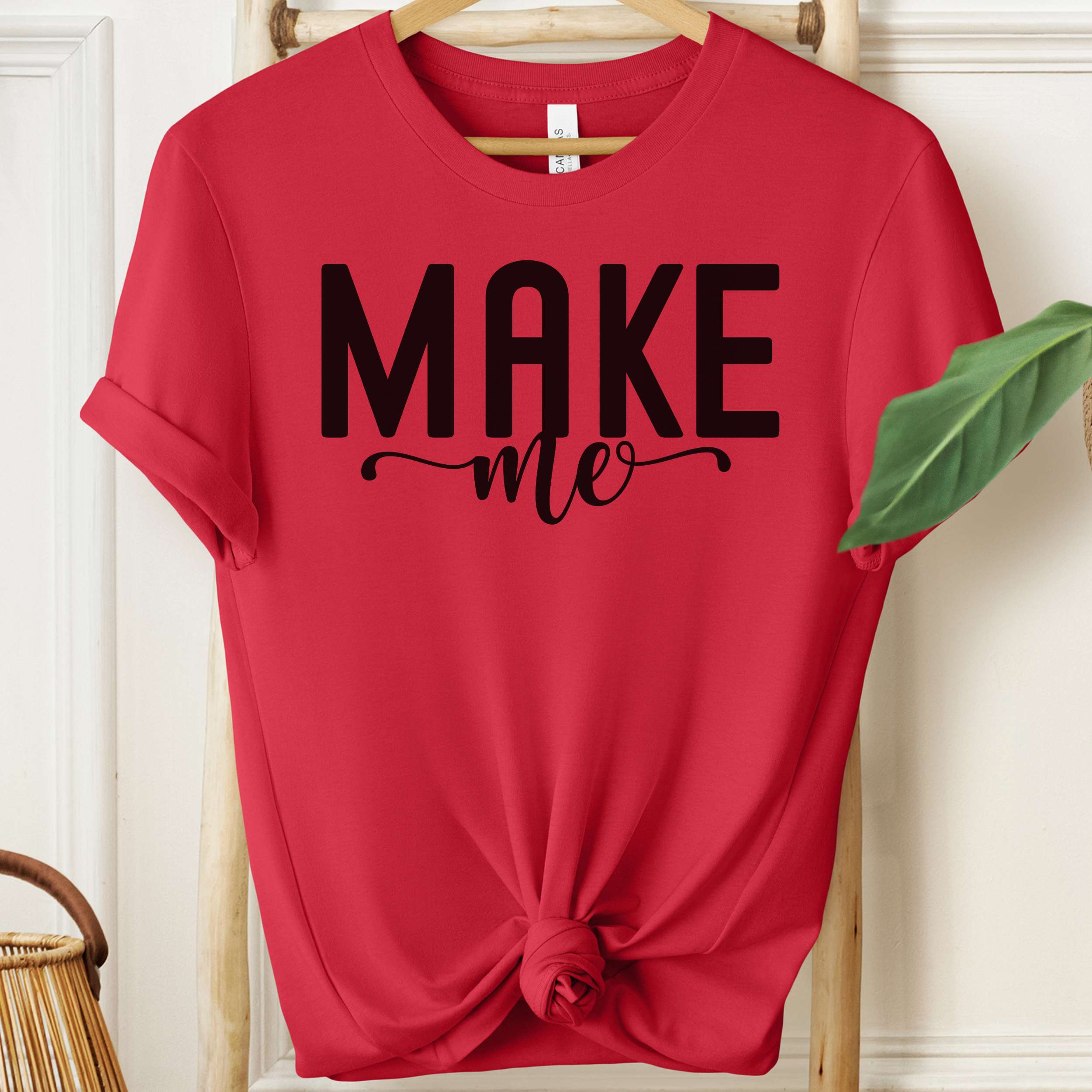 Red t-shirt with 'MAKE me' text on a hanger against a white wall.